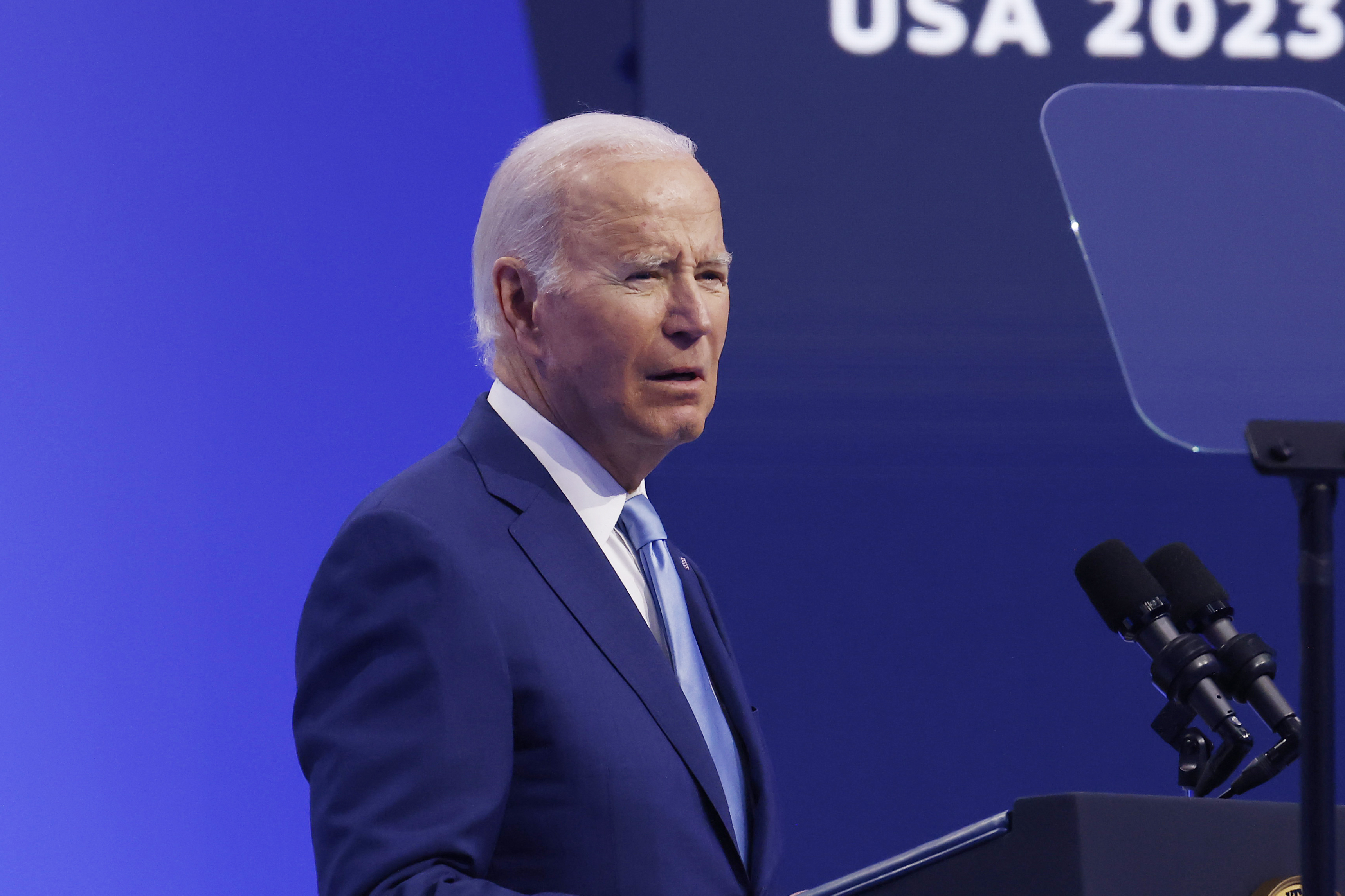 President Joe Biden in a blue suit and light blue tie is speaking at a podium with microphones. The background is blue, and part of the text "USA 2023" is visible.