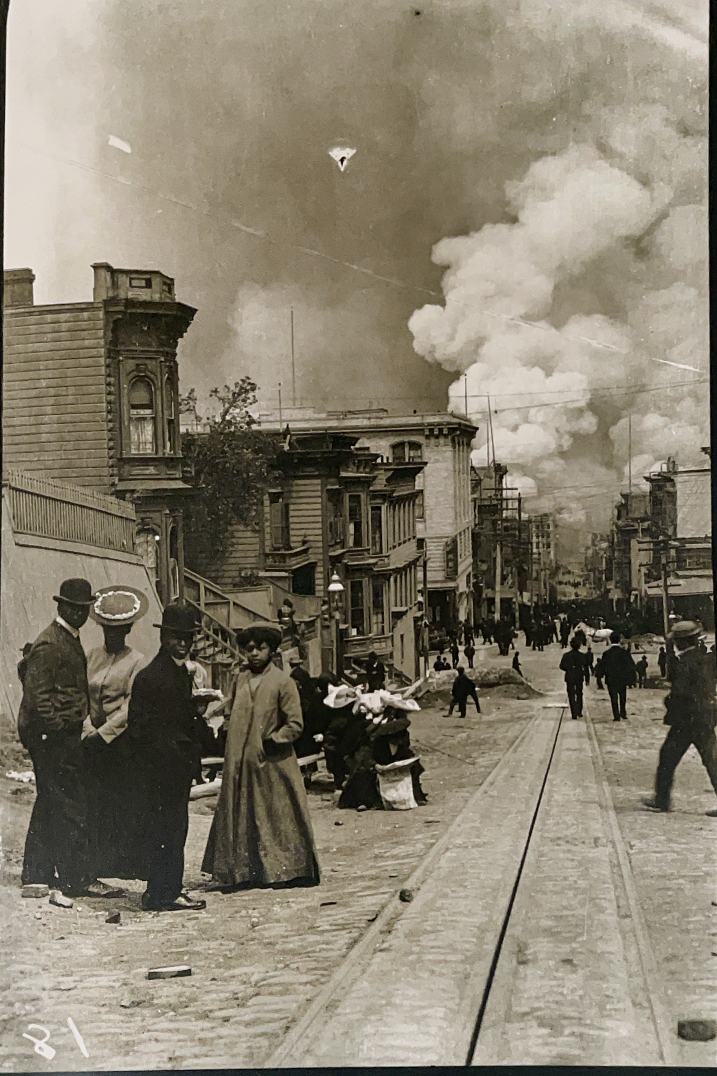 San Francisco residents stand on the street in a black and white photo from 1906, just after the earthquake, with large plumes of smoke in the sky.