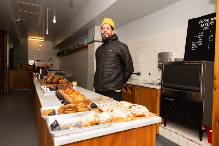A man, Matthew Kosoy poses for a portrait while standing behind a counter at of Rosalind Bakery, with baked goods lining the counter.