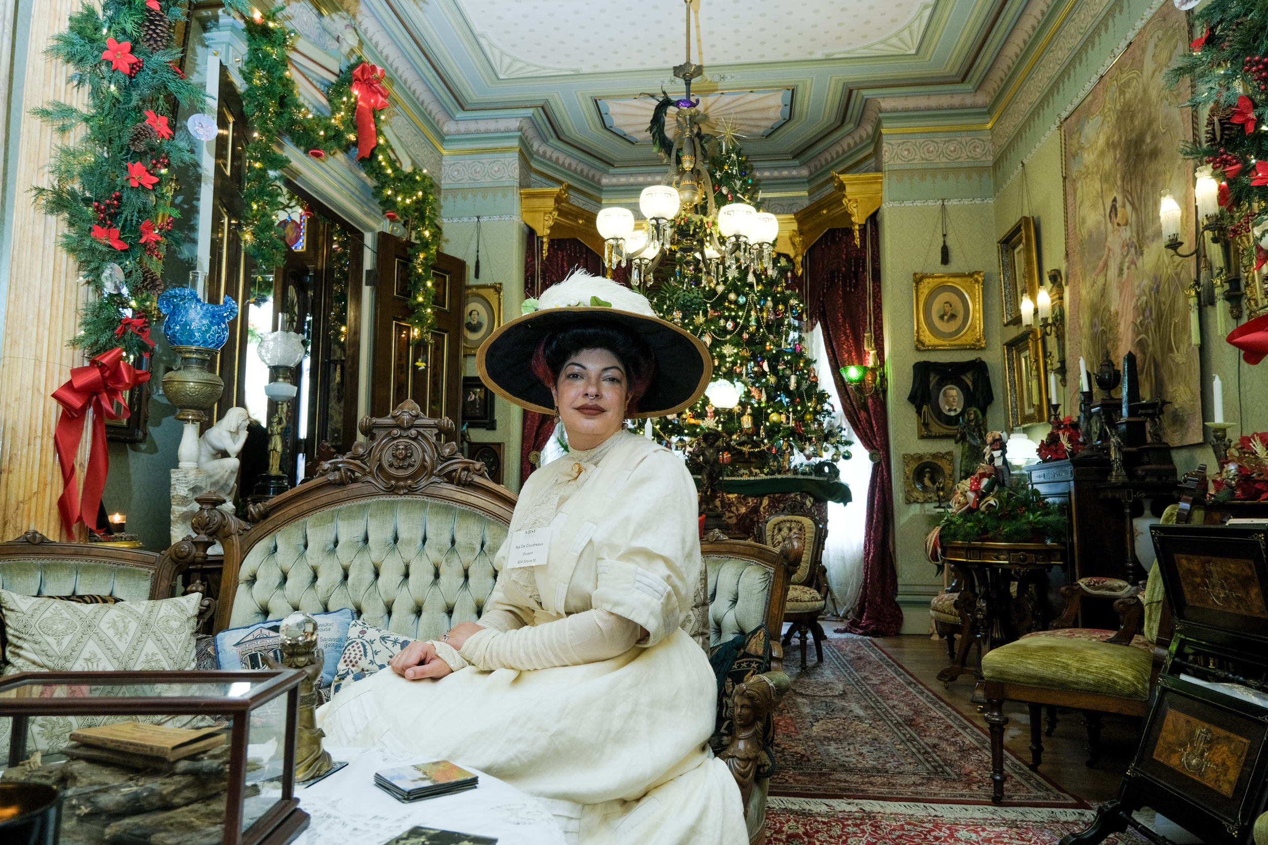 A woman in a white dress and hat sits on a white sofa in a Victorian home filled with antiques.