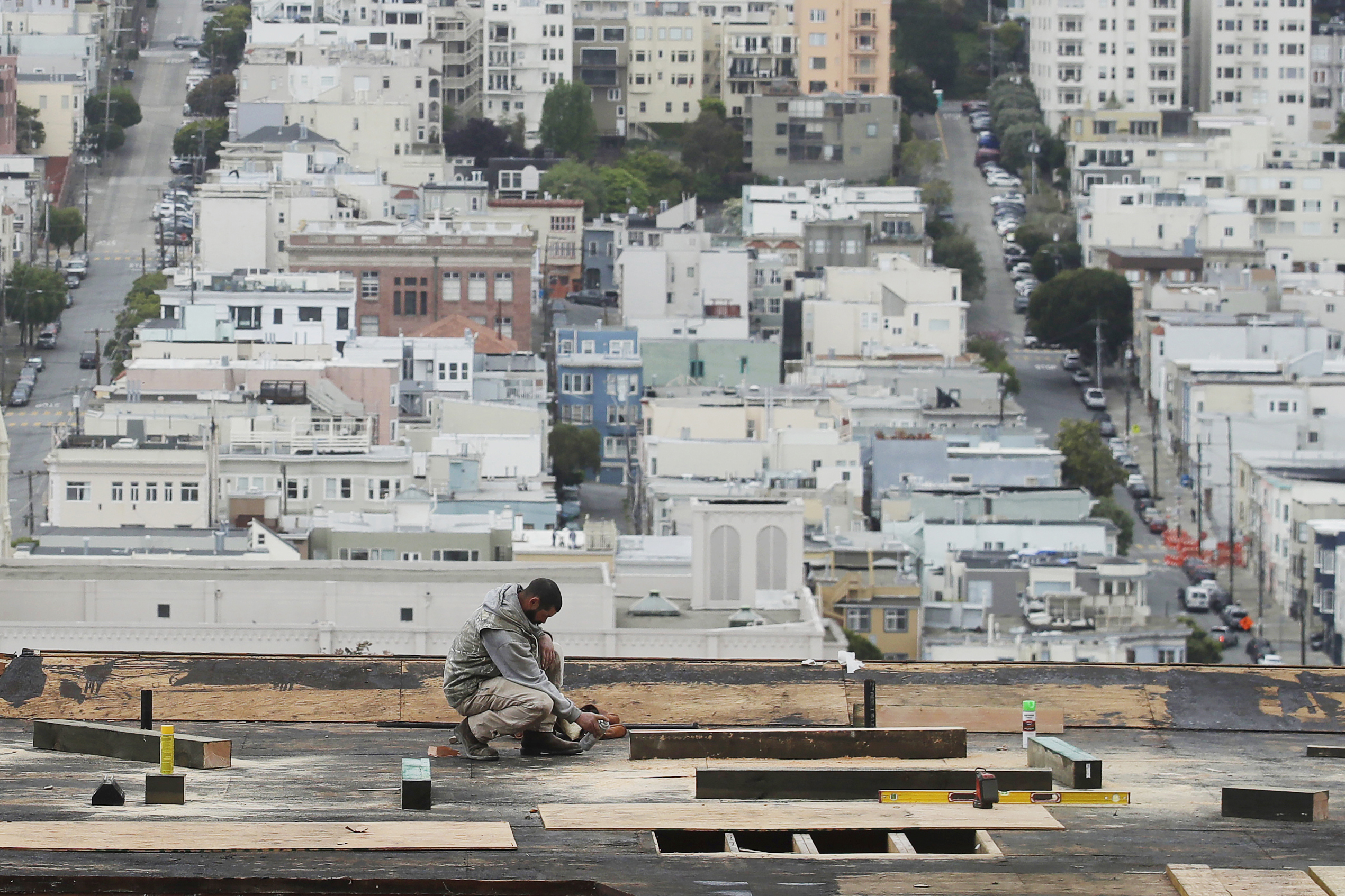 A construction workers works on the roof of a building with a neighborhood with dense homes and housing in the background