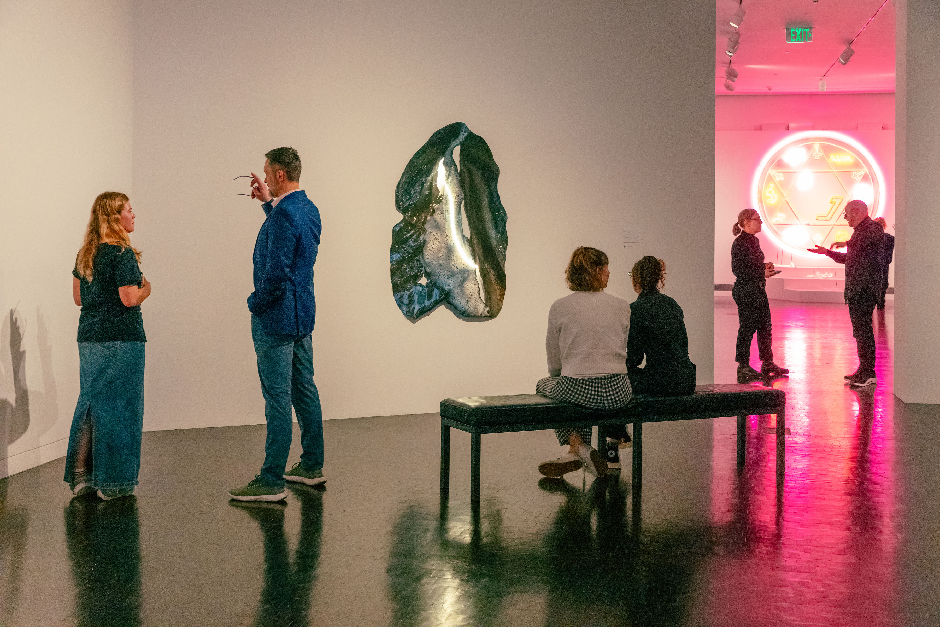 Four people stand and two sit on a bench in a museum exhibition that includes neon artworks glowing pink and white.