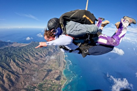 A woman is tandem skydiving, her hand is made into a shaka position suggesting she is having fun, and the background shows the Hawaii coast.