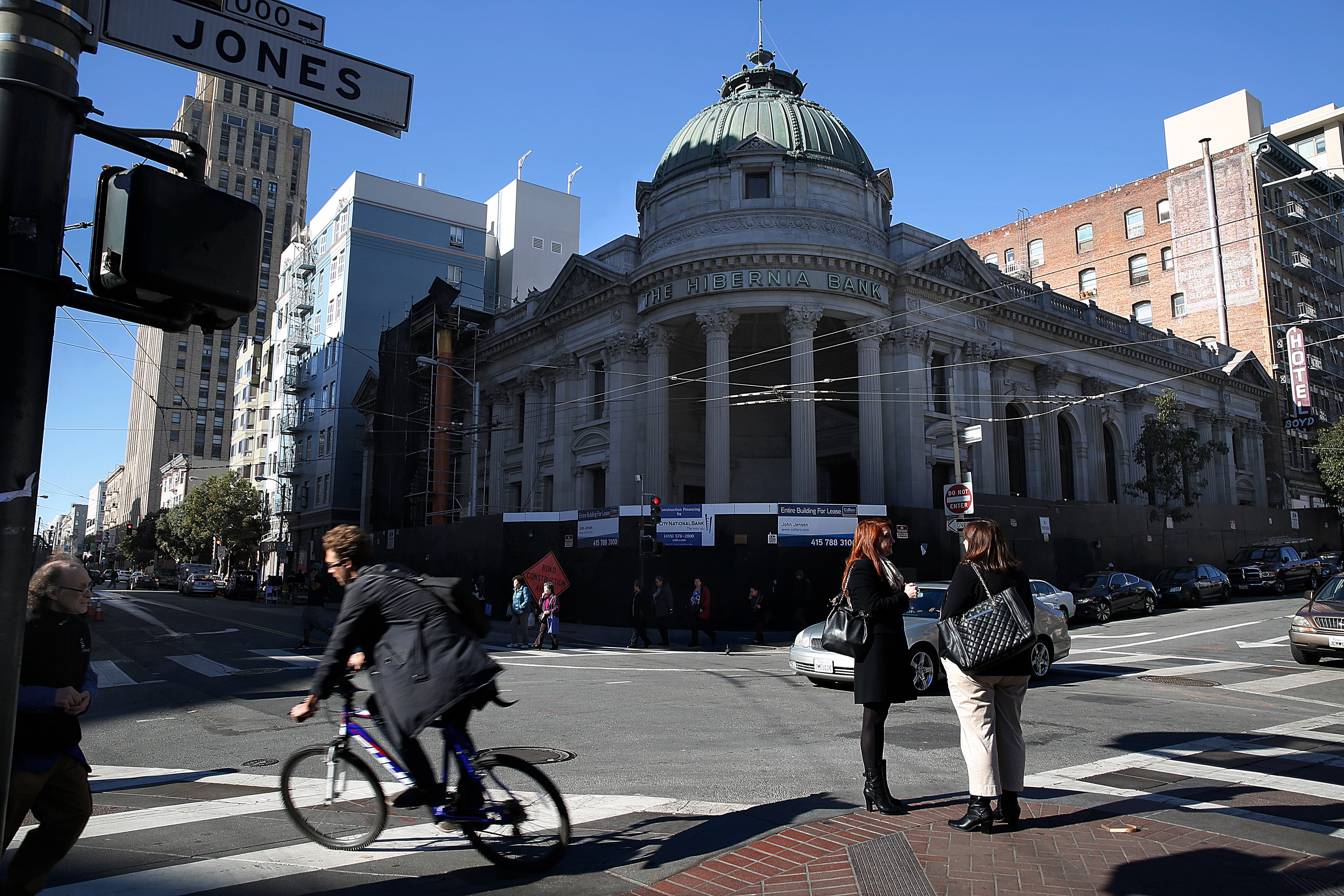 A domed bank building in shadow is seen on a street corner by day with cyclists moving past.