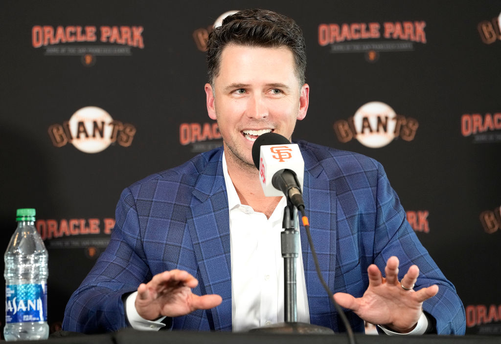 Man in blue blazer speaks in microphone in front of black backdrop advertising the Giants baseball team and Oracle Park