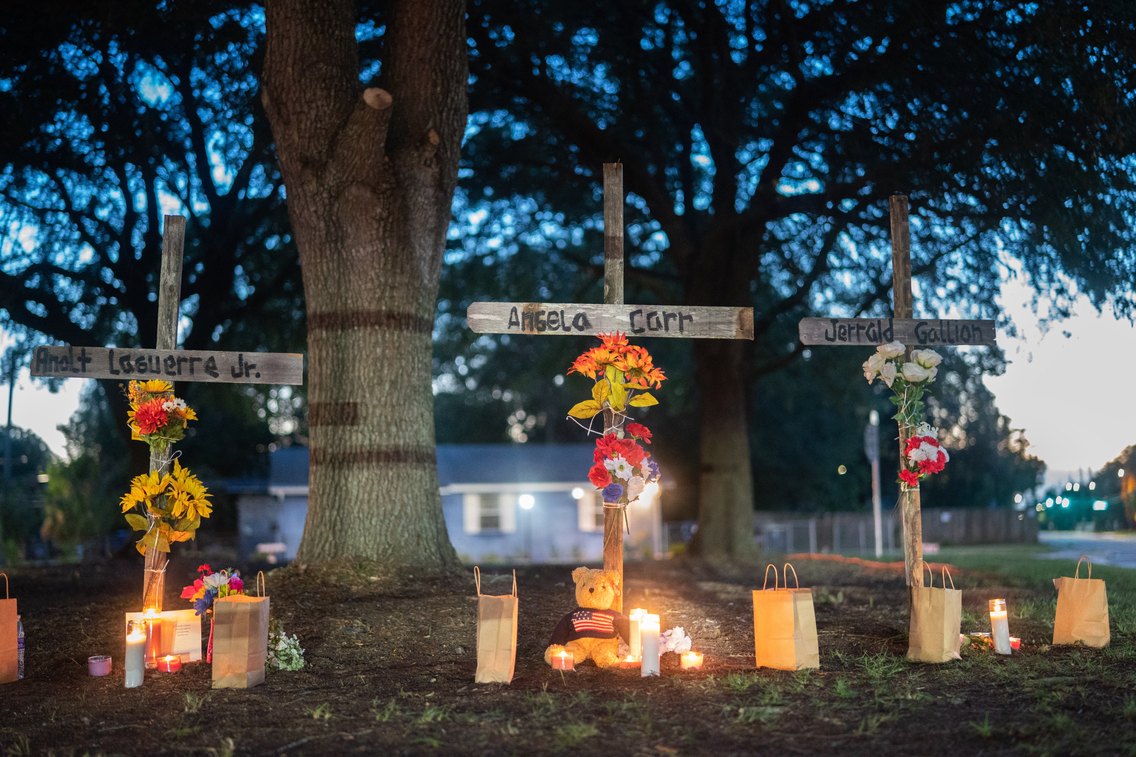 Three grave market crosses stand in front of a tree with flowers tied to them, teddy bears, paper bags and candles are on the ground
