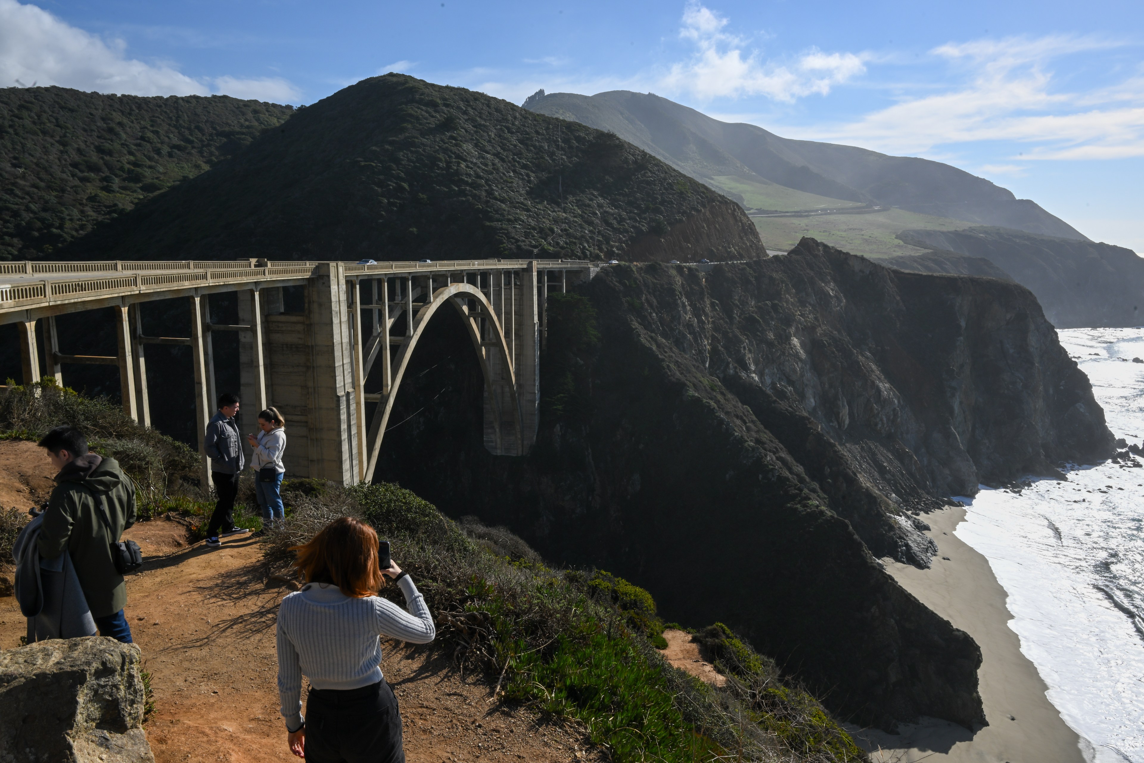 Bixby Bridge in the background and people in the foreground taking pictures and posing right next to the clif.