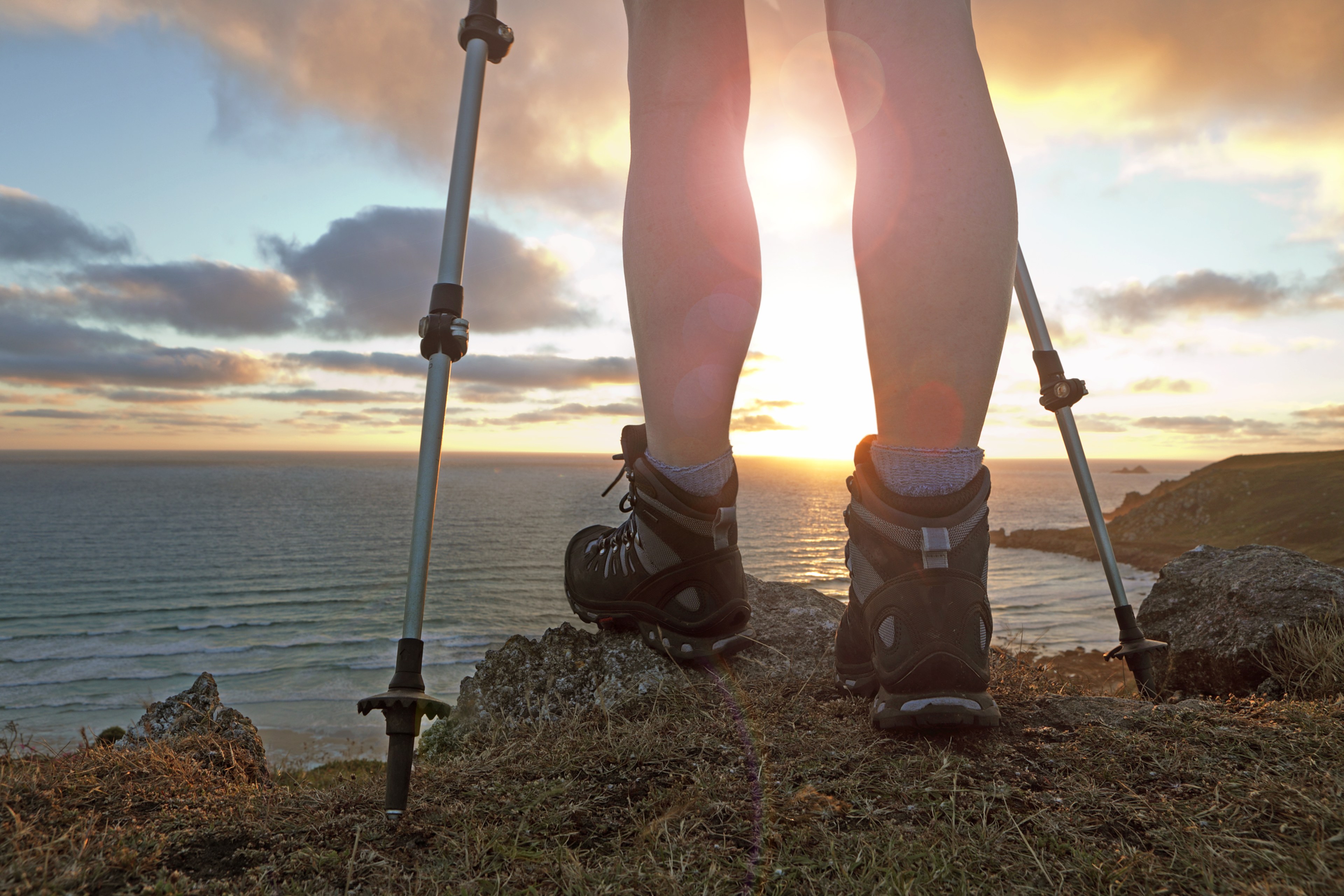 A person with hiking poles and boots stands at an edge of a cliff looking out into the ocean. 
