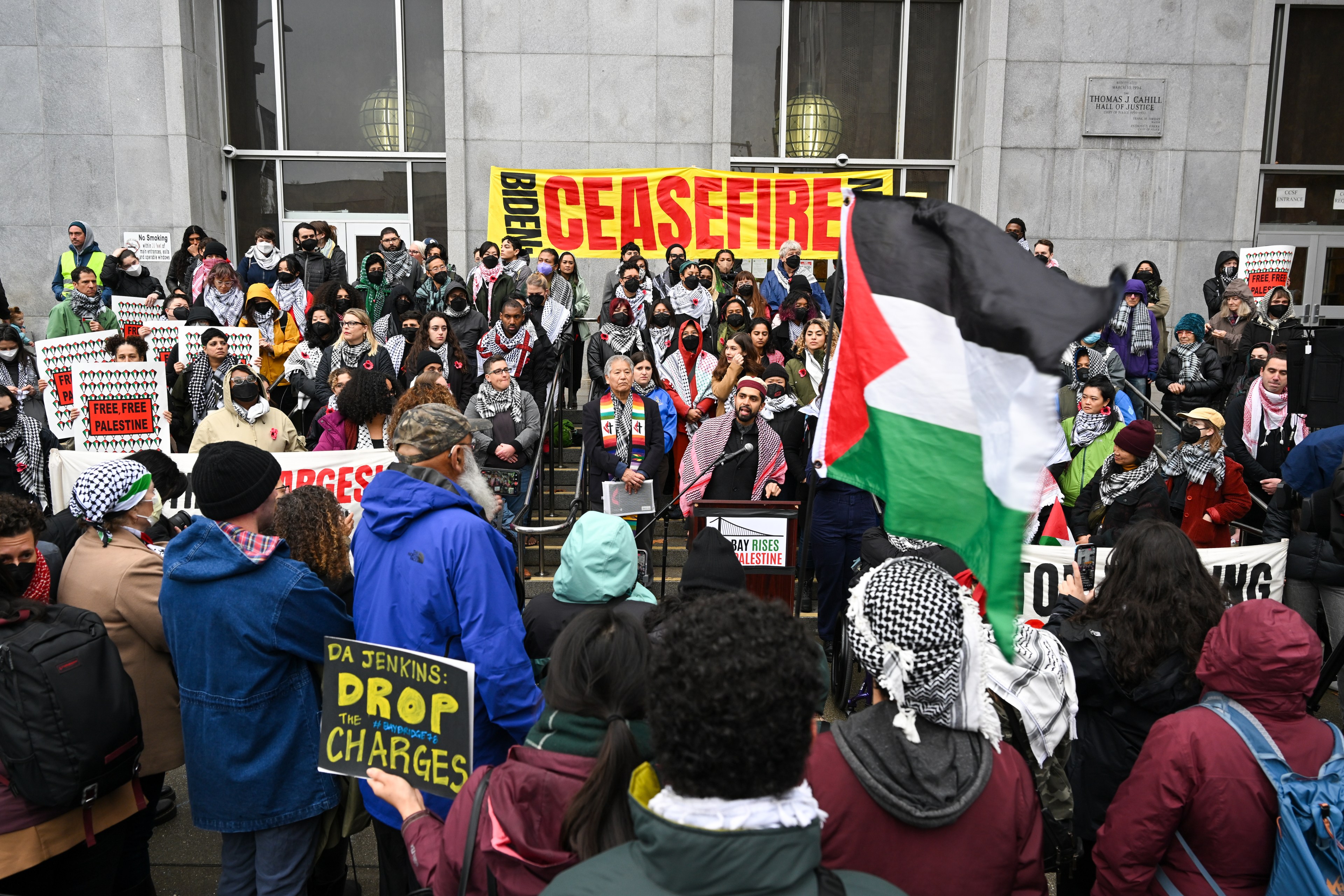 A crowd gathers on steps Superior Court in San Francisco with banners that read "BIDEN CEASEFIRE", "DA JENKINS DROP THE CHARGES" along with a Palestinian flag waving in the foreground.