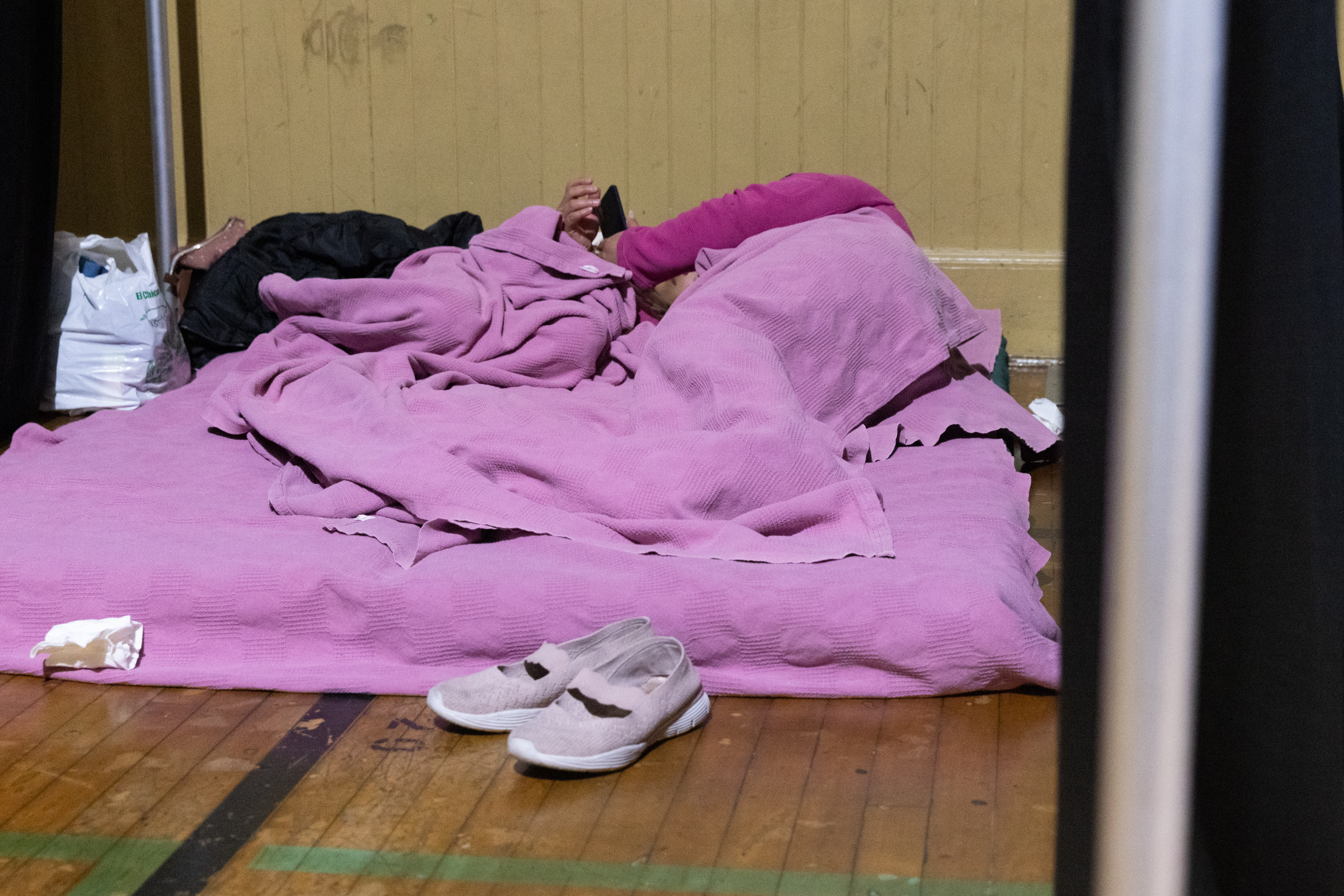 Two people curl up under a pink blanket on a mattress on the floor.