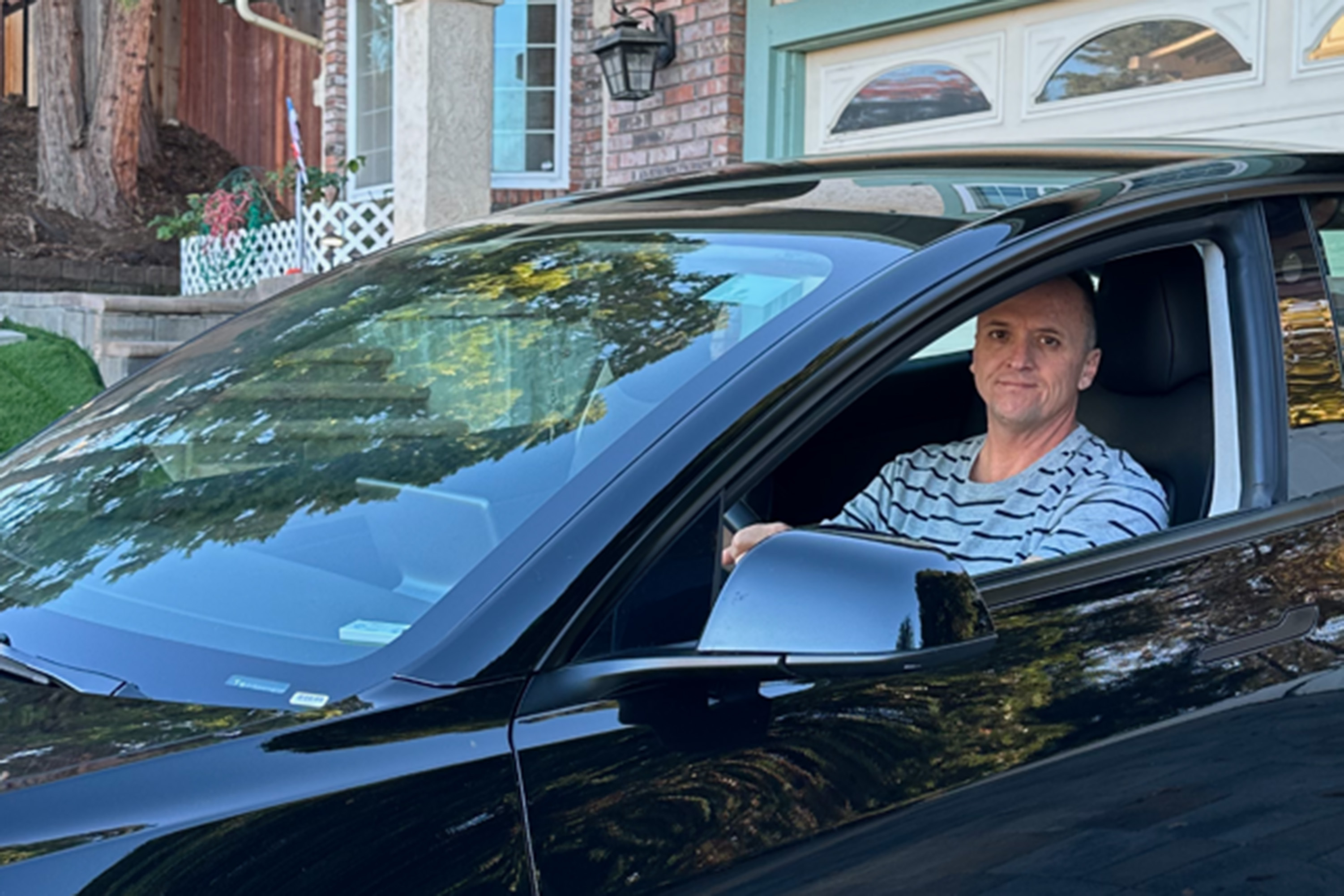 A man sits inside a black sedan parked in a driveway.