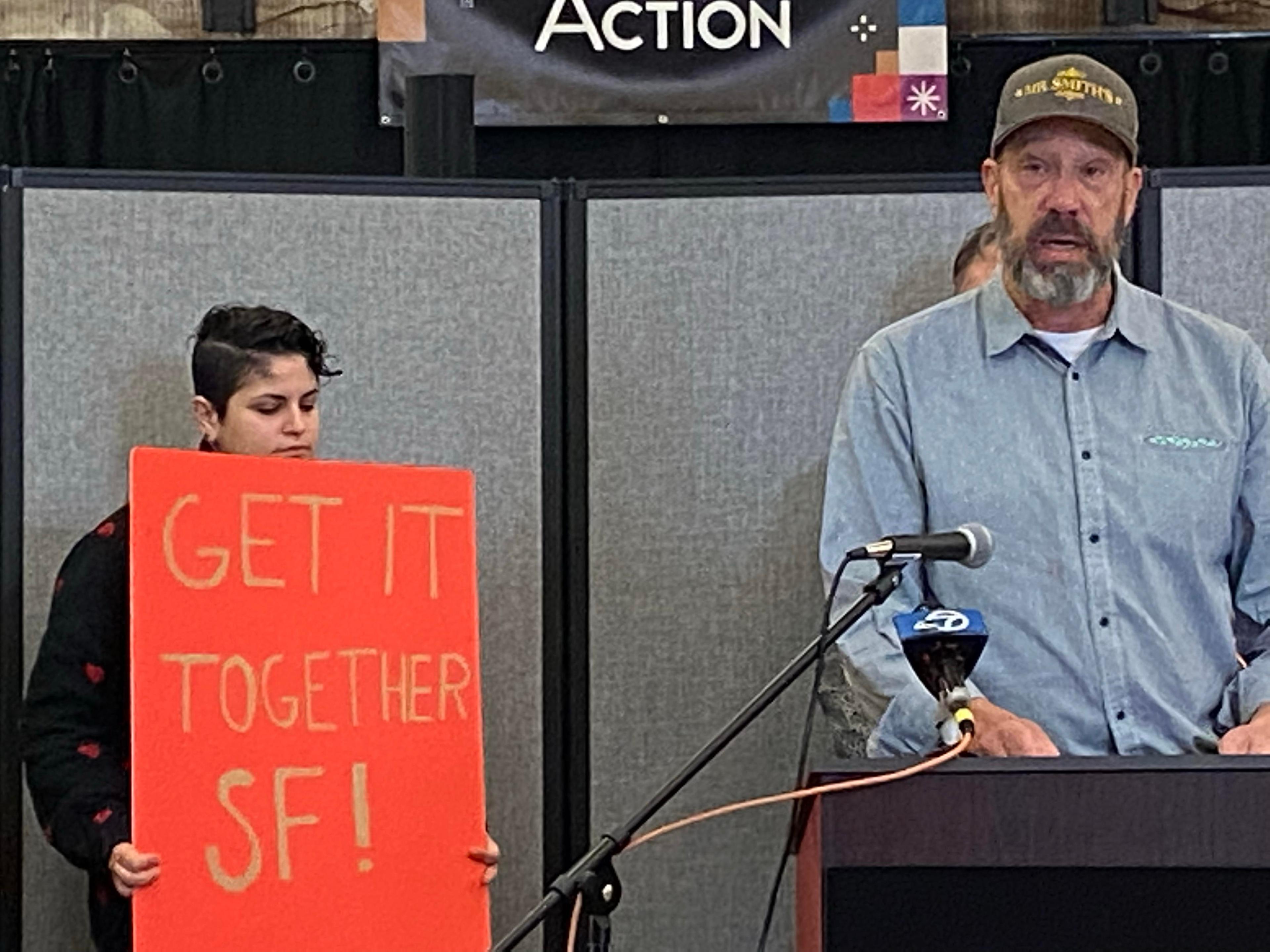 Max Young, a bar owner, stands behind a podium next to a women with a sign reading &quot;get it together SF.&quot;
