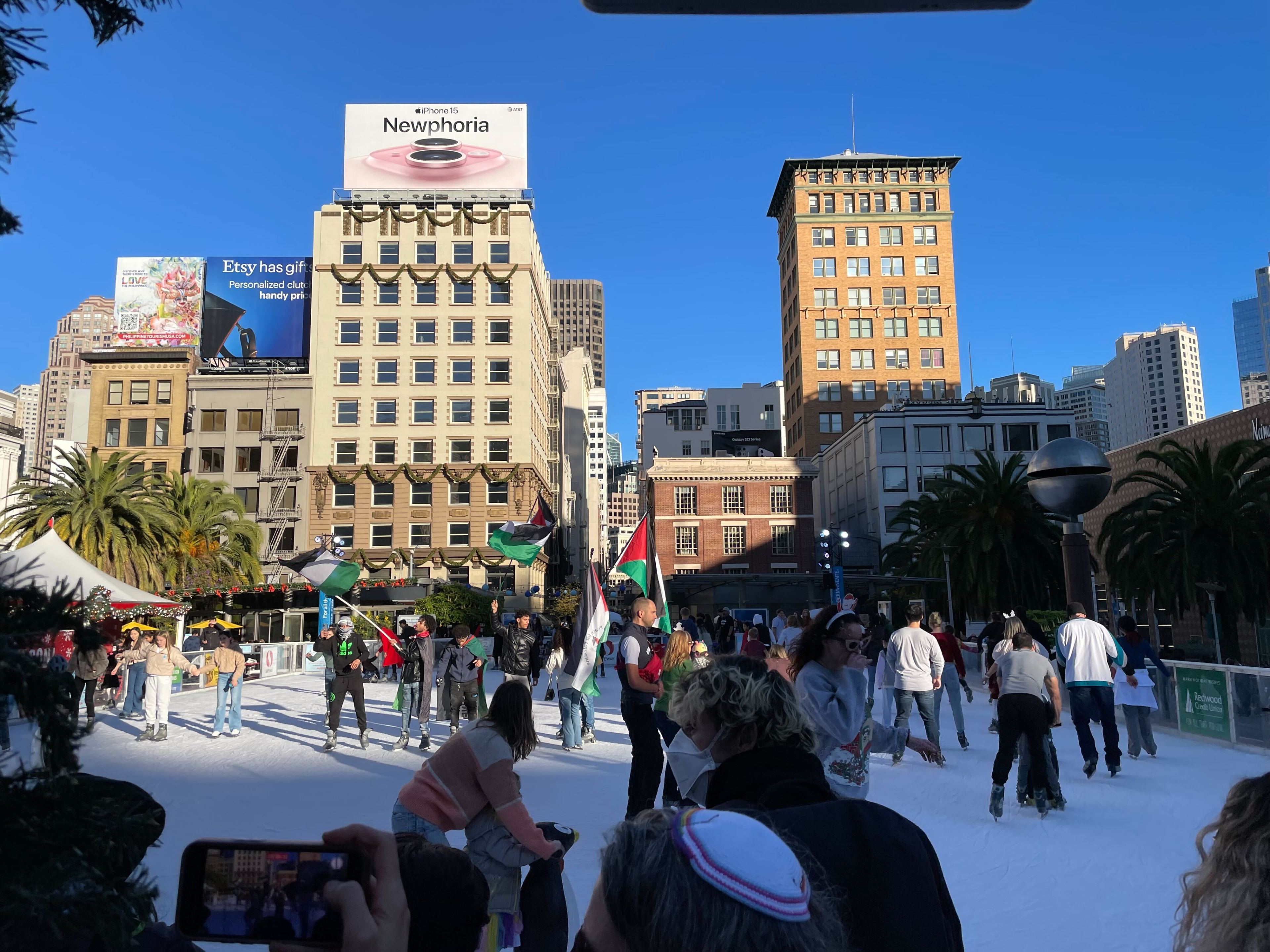An outdoor ice rink bustling with skaters, surrounded by palm trees, with tall buildings and ads overhead.