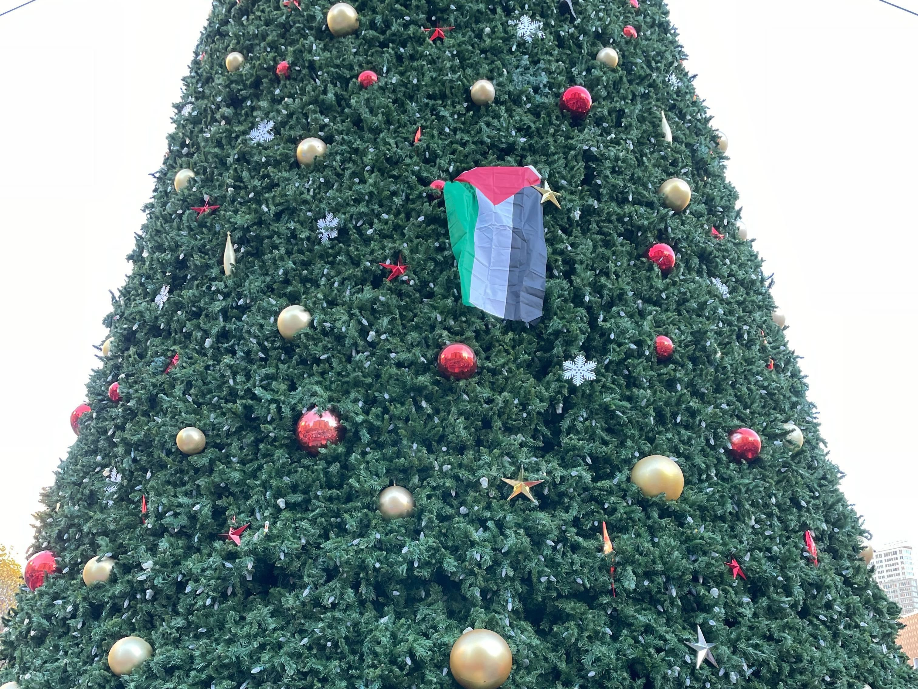A large Christmas tree decorated with red and gold ornaments, and a Palestinian flag in its branches.