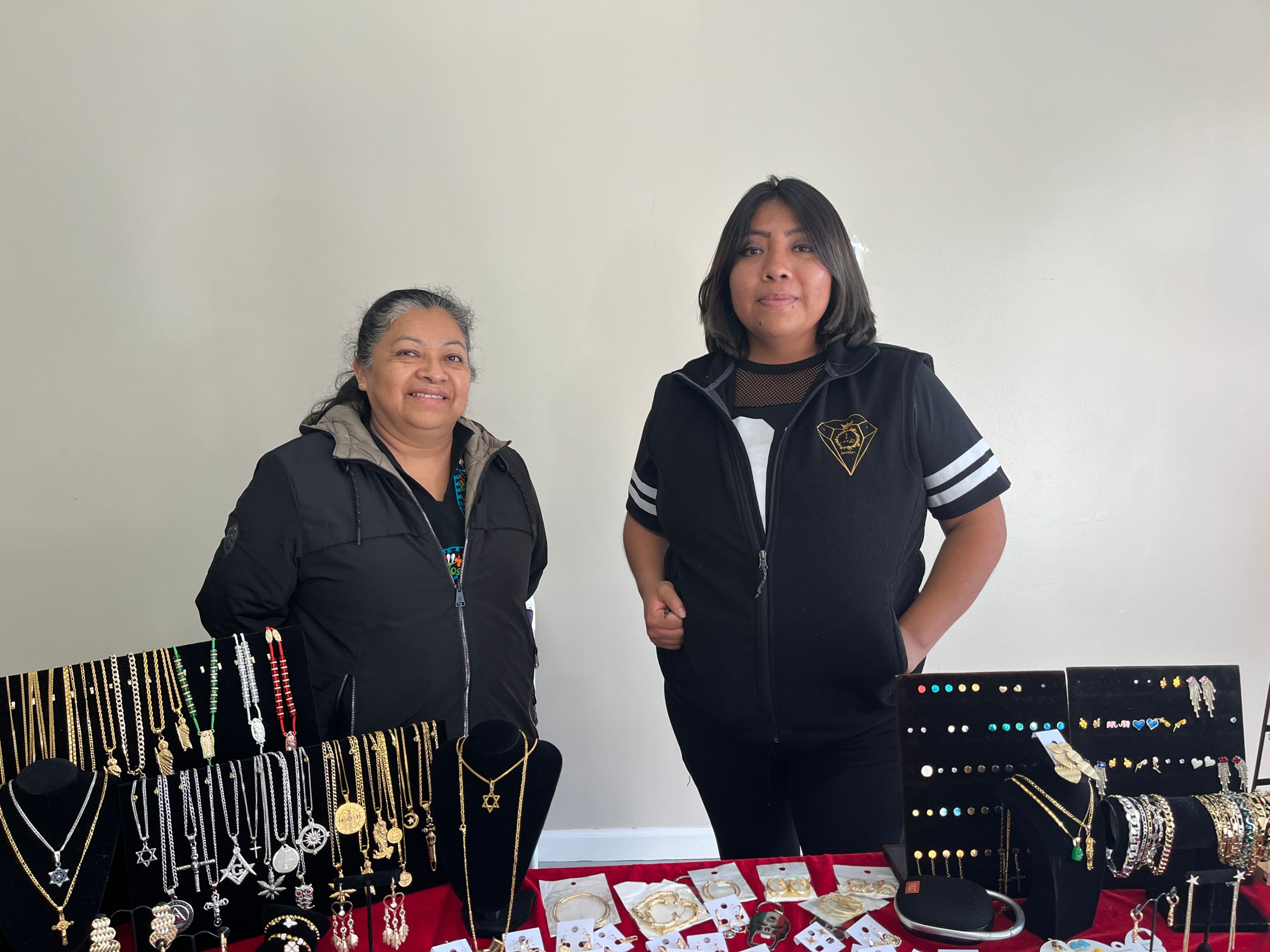 An older woman stands next to a younger woman behind a table with silver and gold jewelry