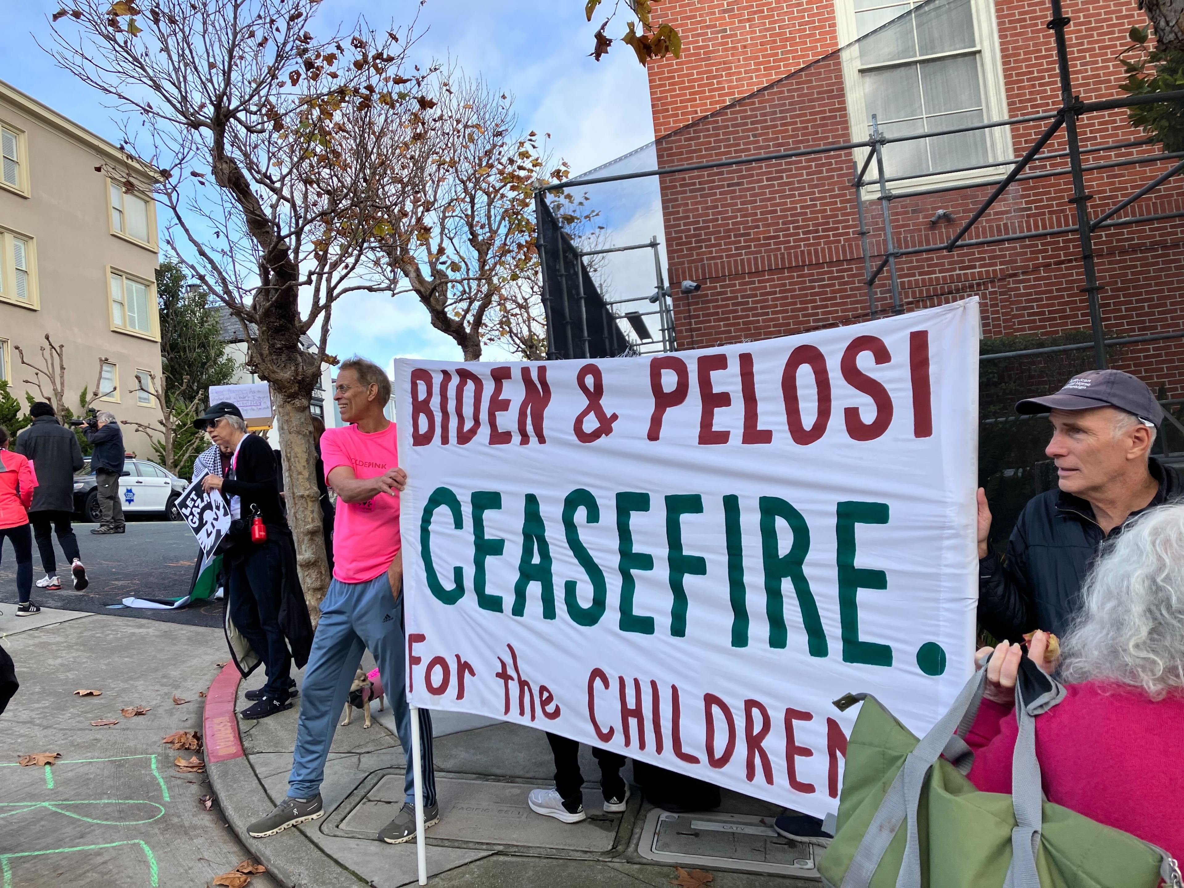People gather on a street corner with sign that says Biden and Pelosi ceasefire for the children