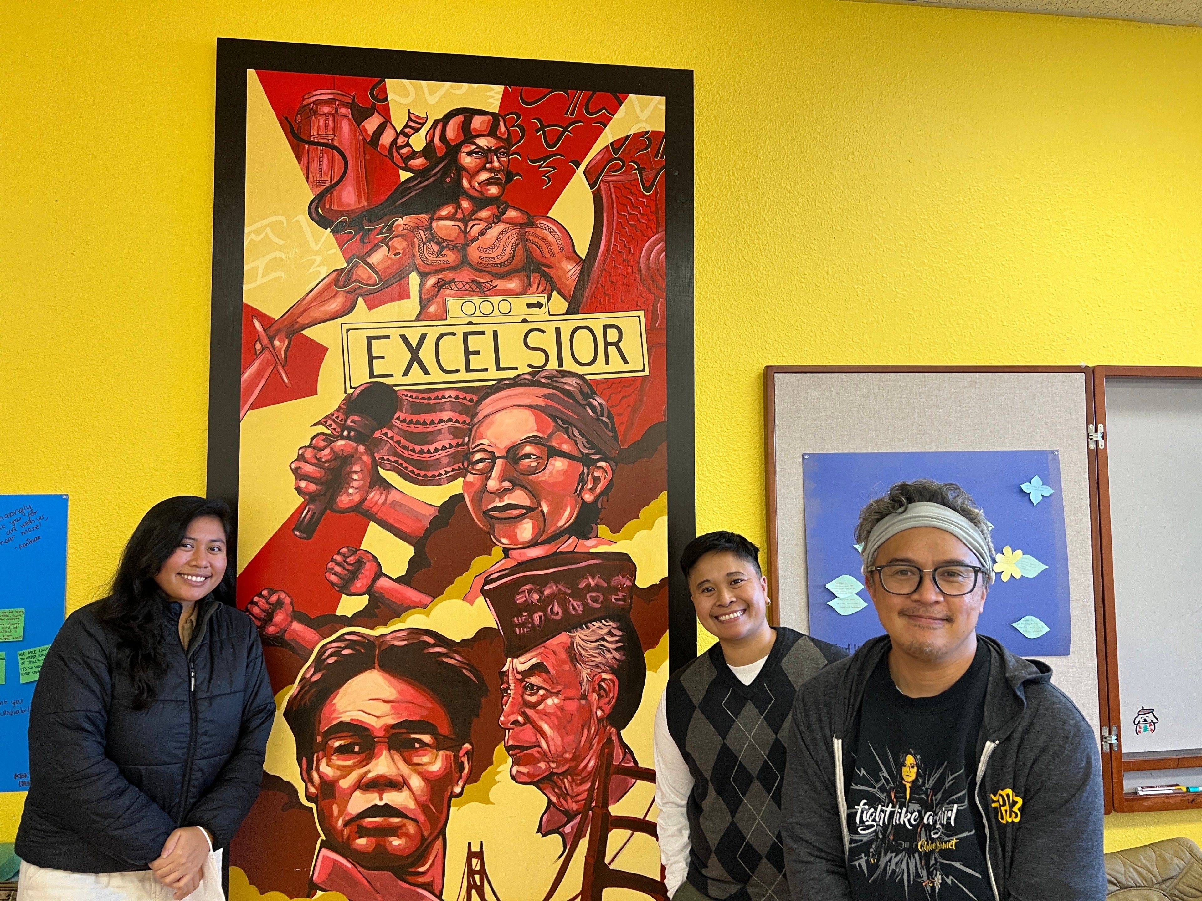 Three people stand in front of a red and yellow mural that reads &quot;Excelsior.&quot;
