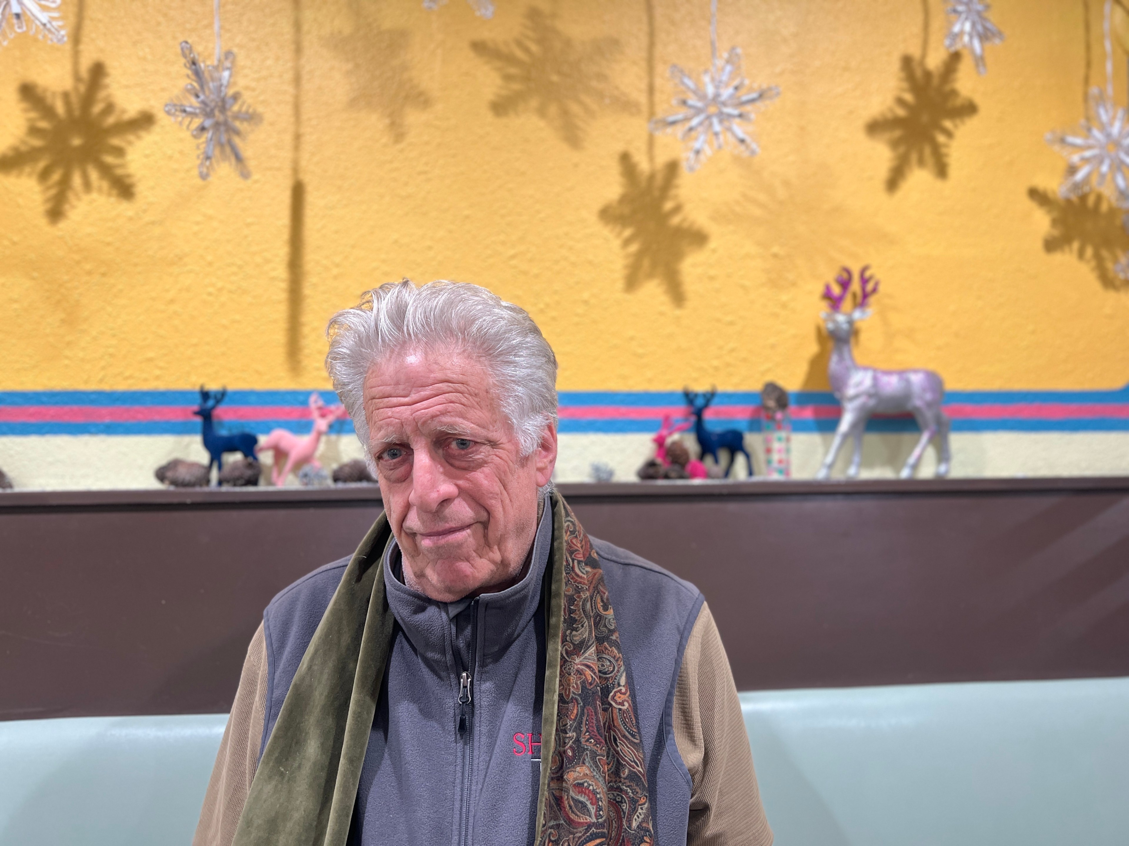 A man with white hair and a blue vest sits in a booth with reindeer and snowflakes behind him.