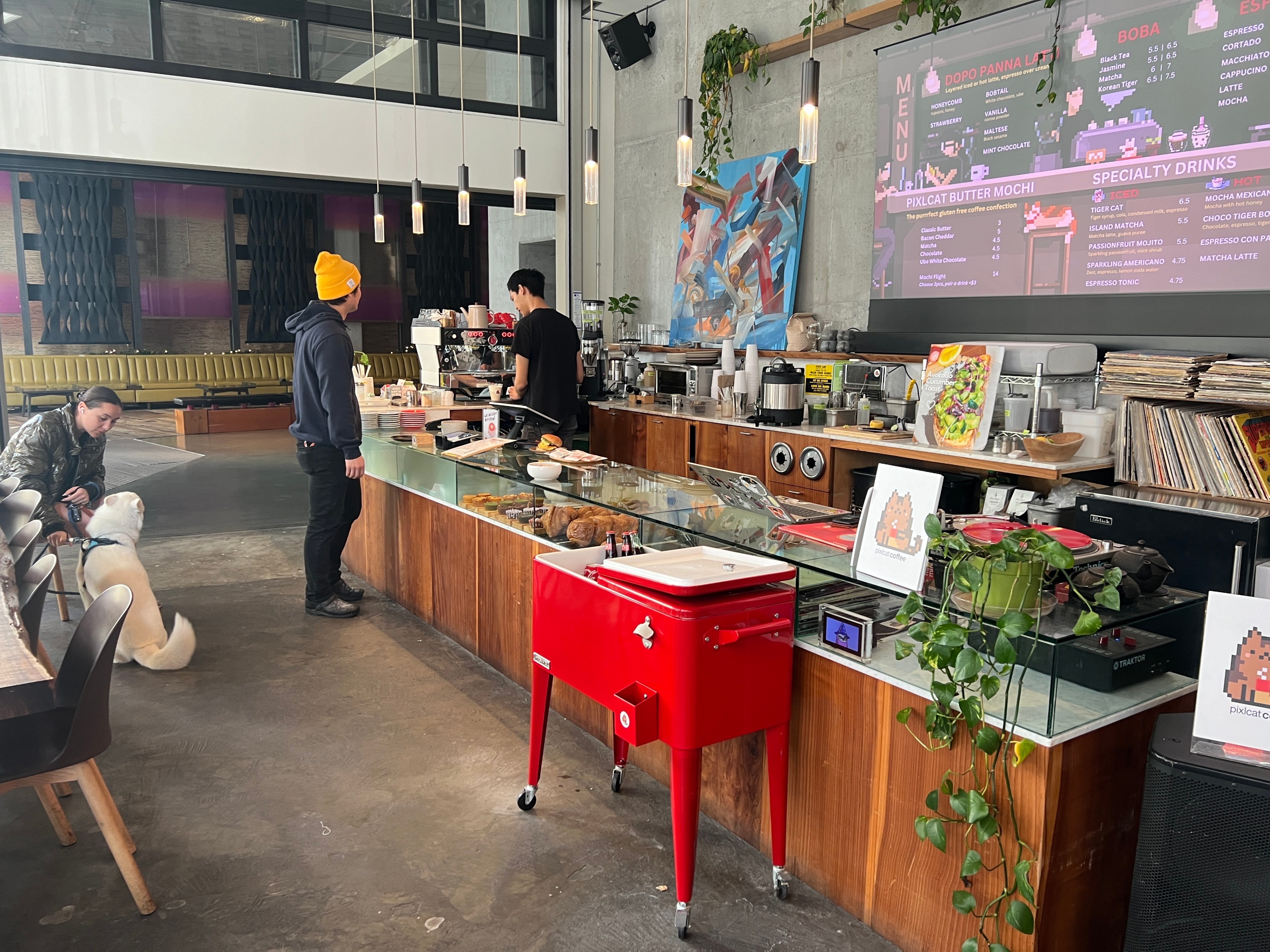 A long wooden bar stands inside a coffee shop with a menu screen behind it. 