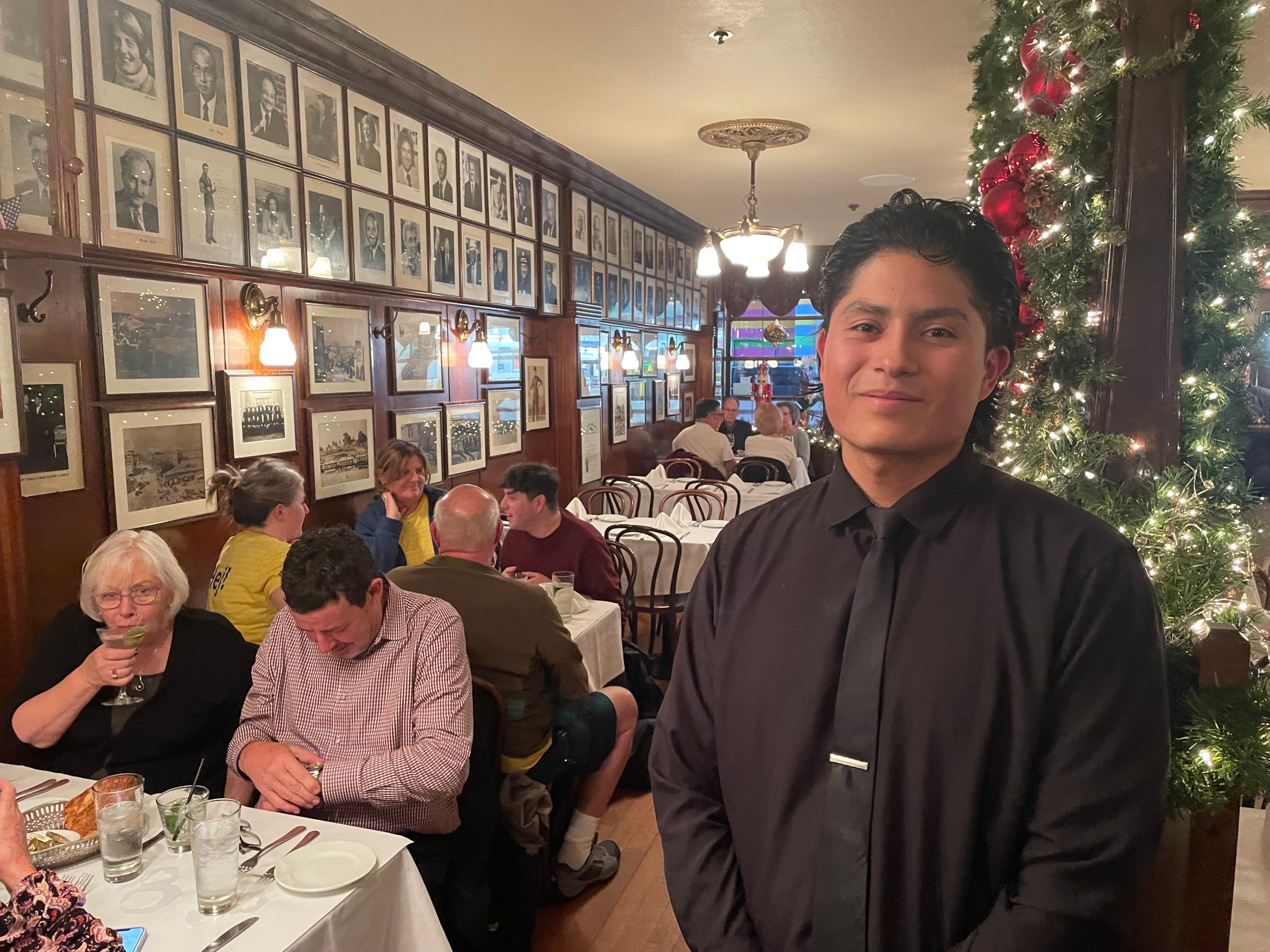 A waiter poses for a portrait in front of restaurant tables filled with customers.