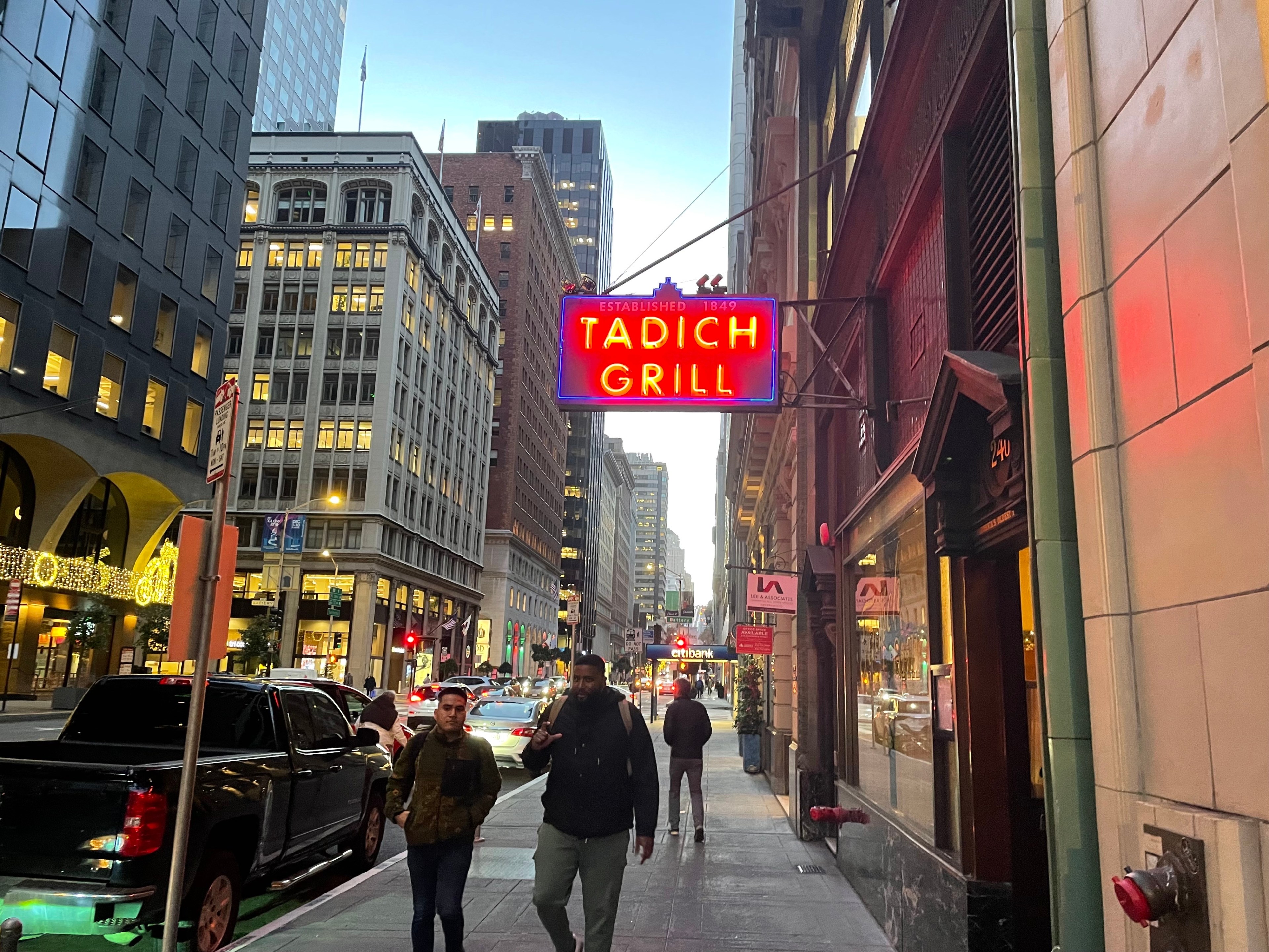 Pedestrians walk under a restaurant's neon blade sign overhanging a city sidewalk.