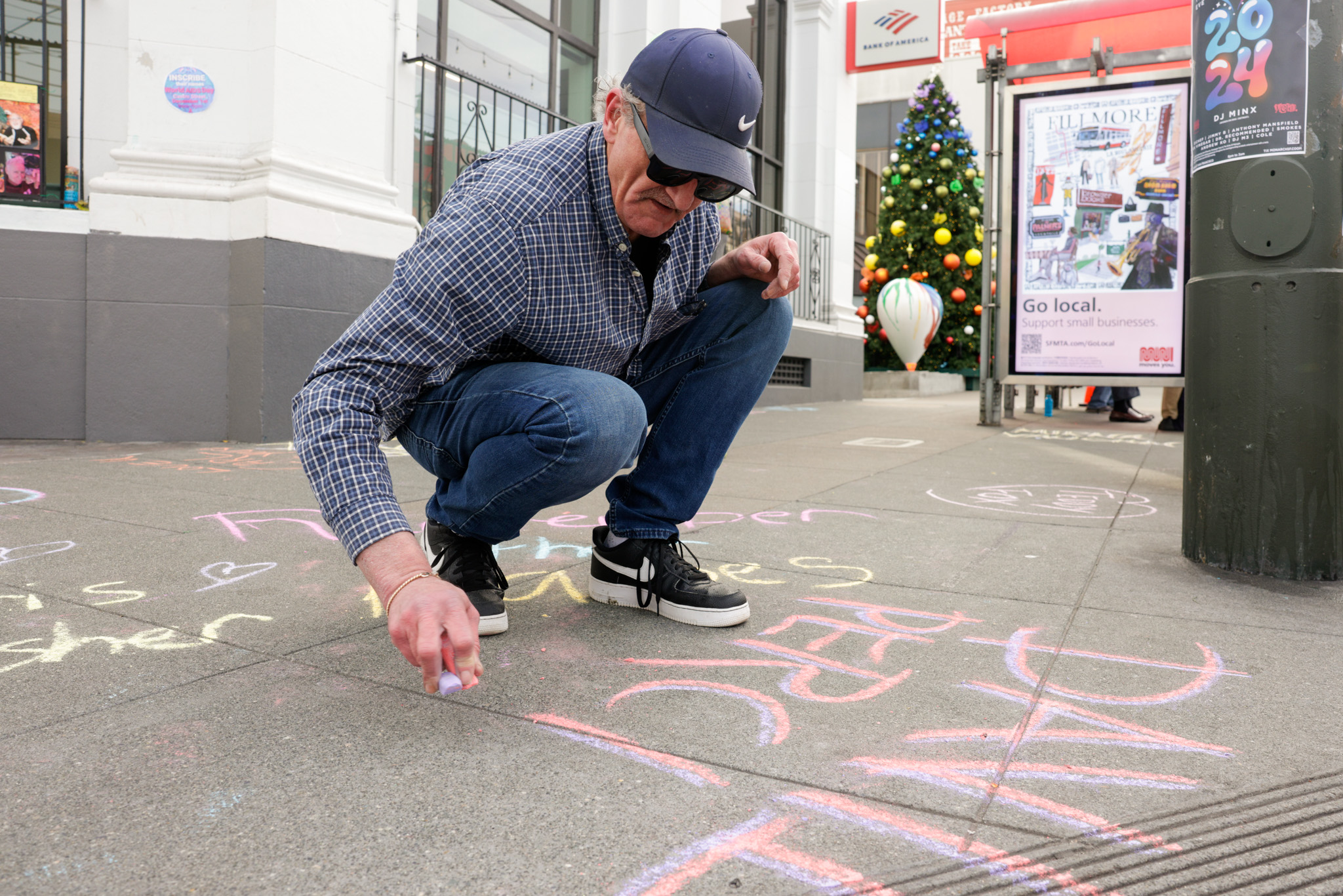 A person in a blue baseball cap writes a person name in pink and purple chalk during World Aids Day.