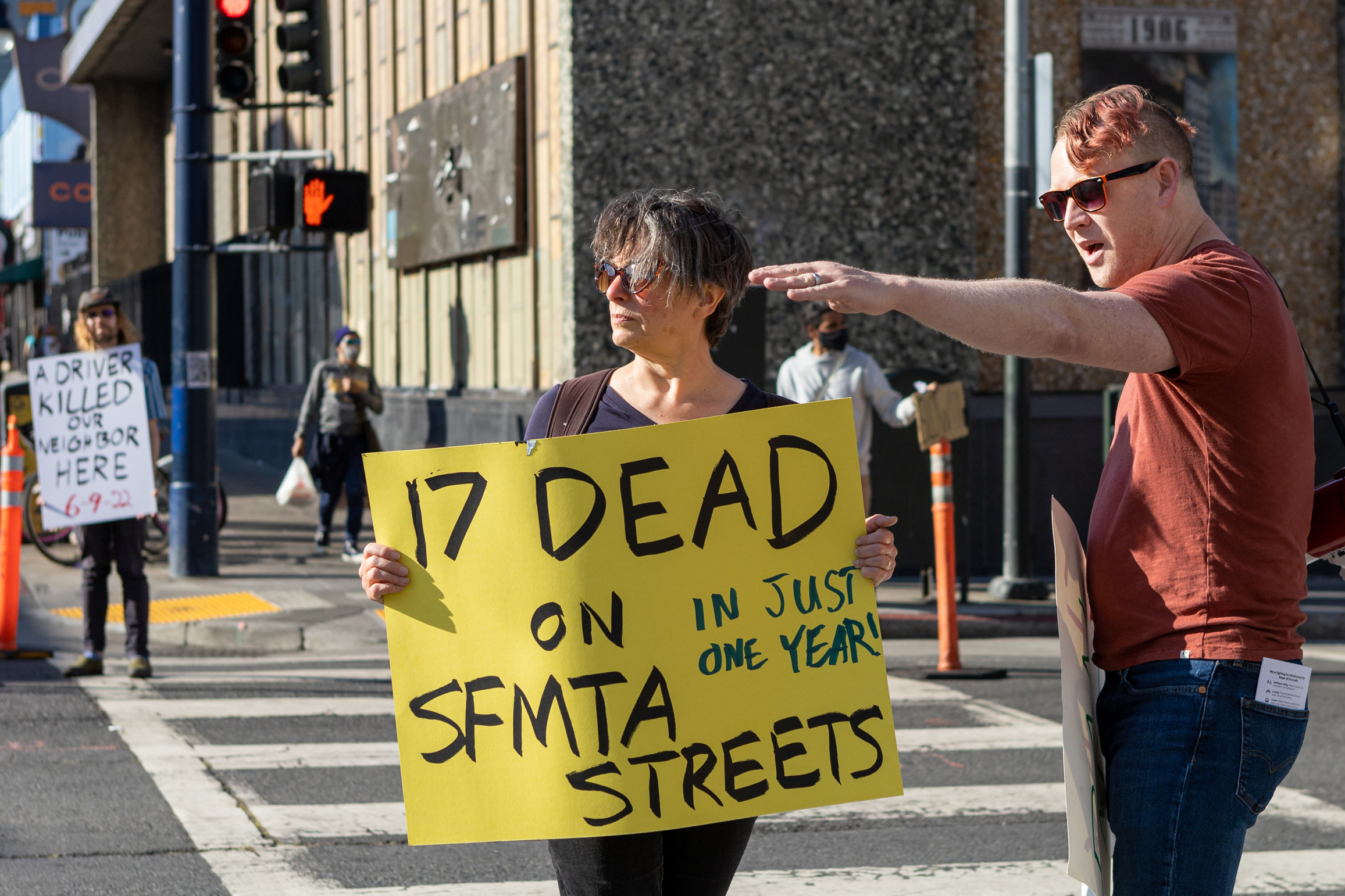 Two people stand on a city street and one person holds a sign that reads &quot;17 DEAD ON SFMA STREETS IN JUST ONE YEAR&quot;