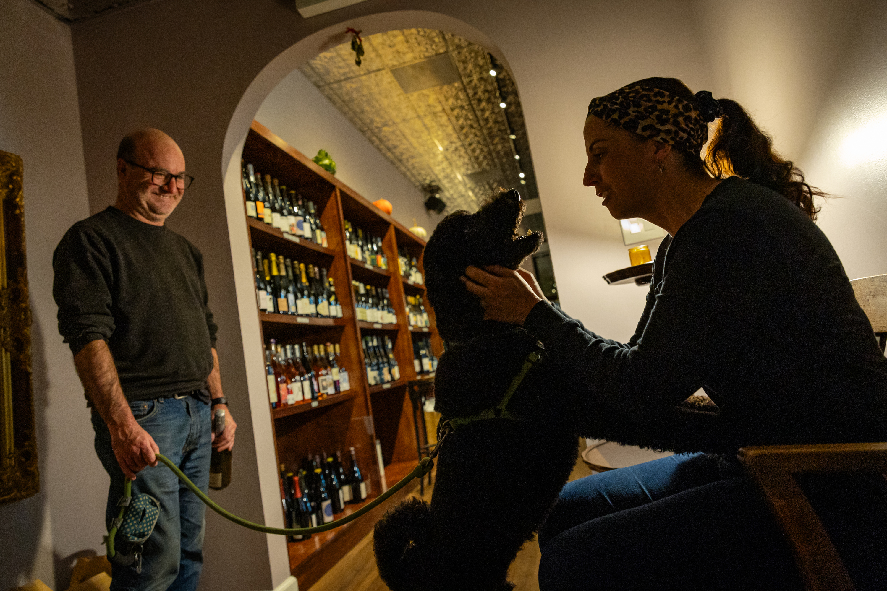 A woman pets a dog in a wine shop as the dog owner smiles.