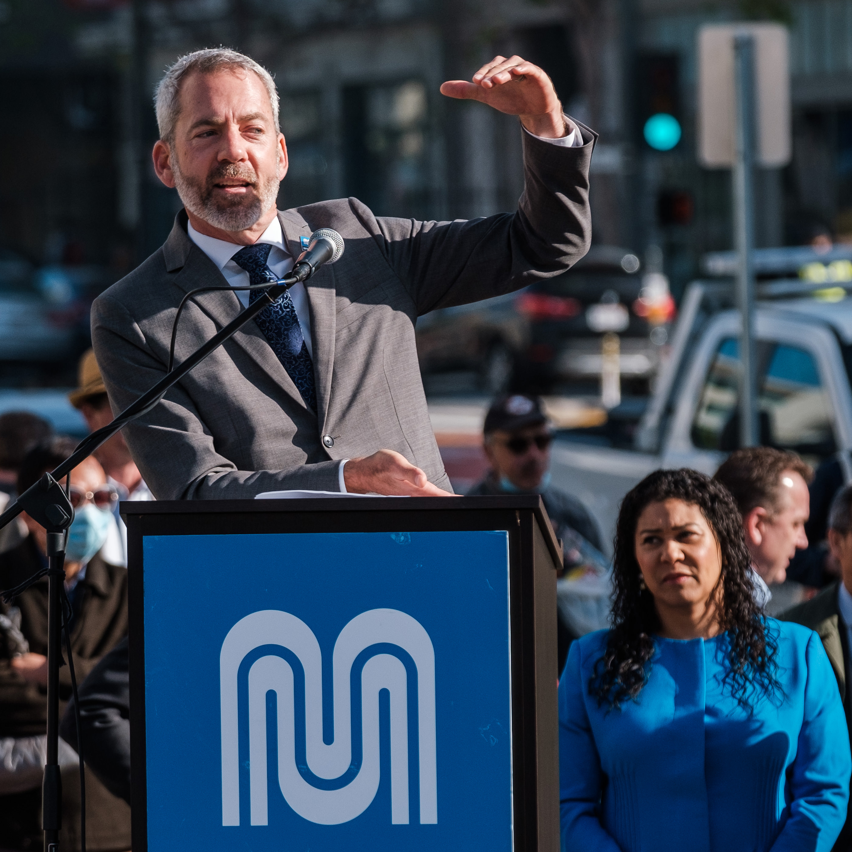 Jeffrey Tumlin the San Francisco Director of Transportation raises his arm while giving a presentation with mayor london breed in the background.
