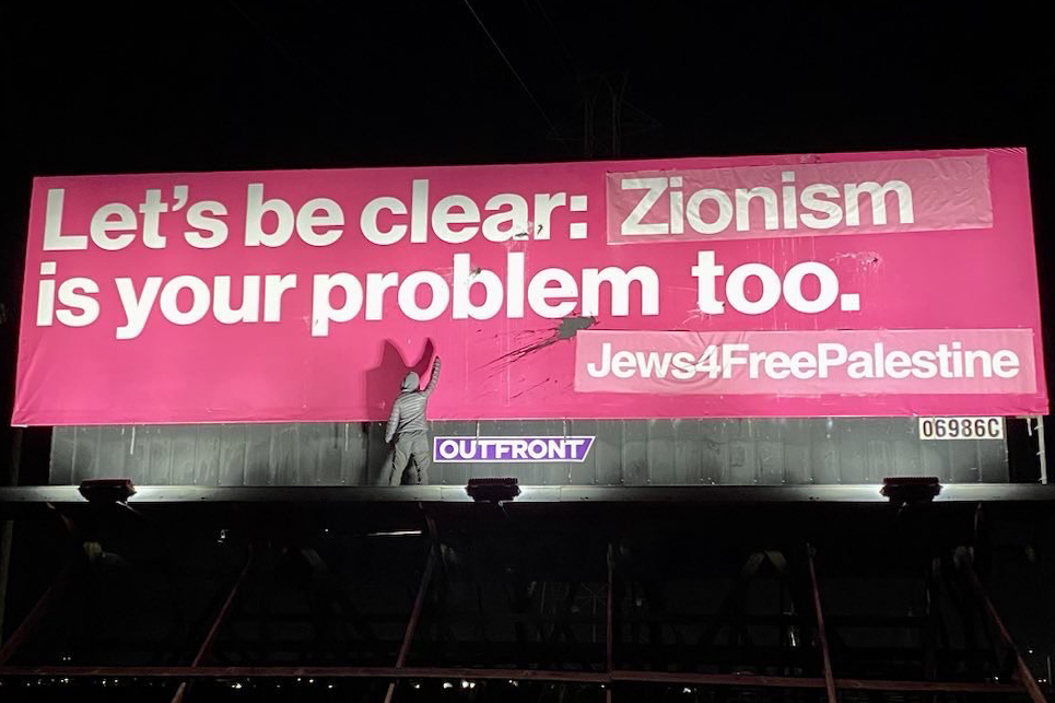 A person raises their fist while standing below a billboard that reads “Lets be clear: Zionism is your problem too. Jews4FreePalestine” in the evening.