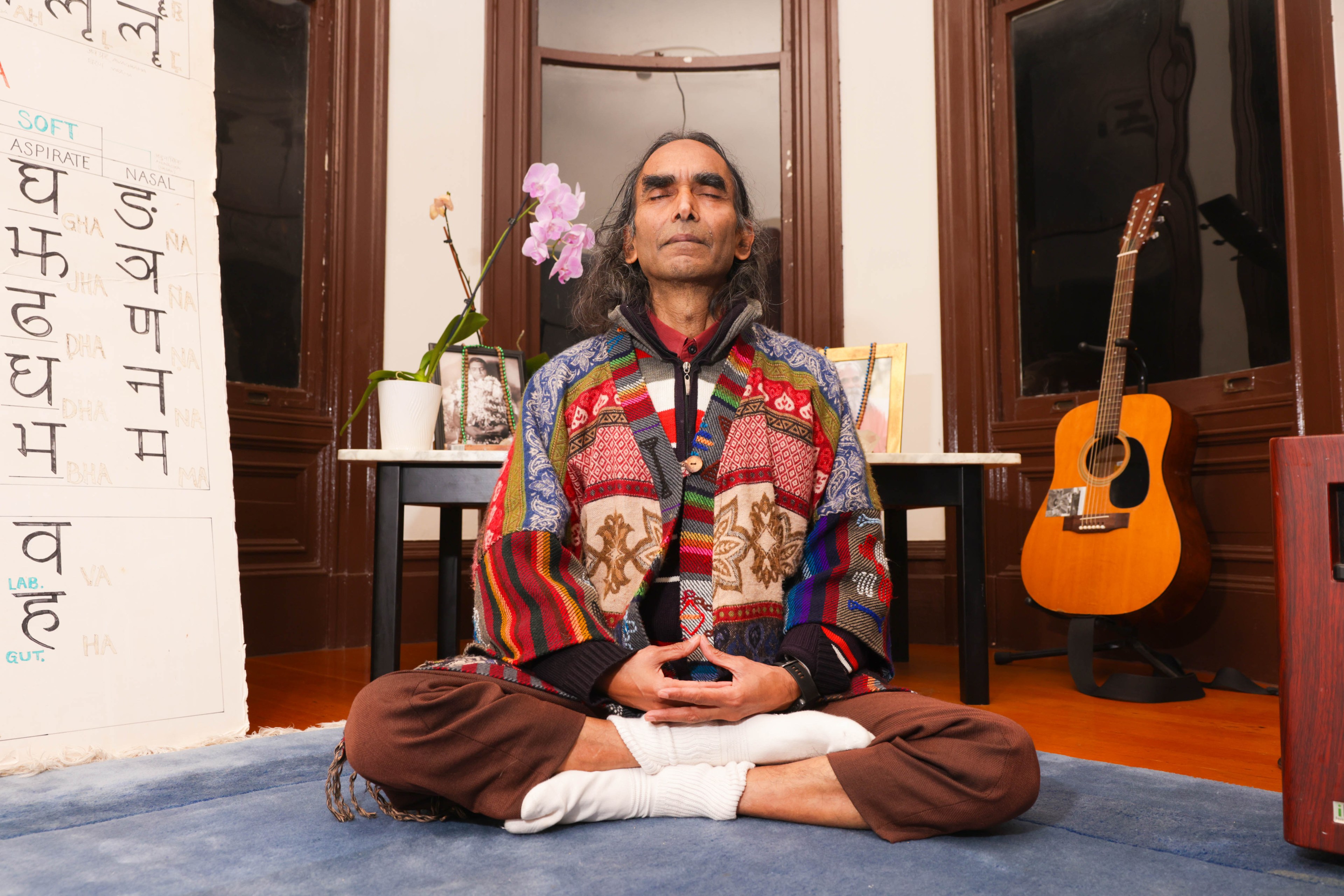 A man sitting in a yoga pose with a guitar and Sanskrit in the background.