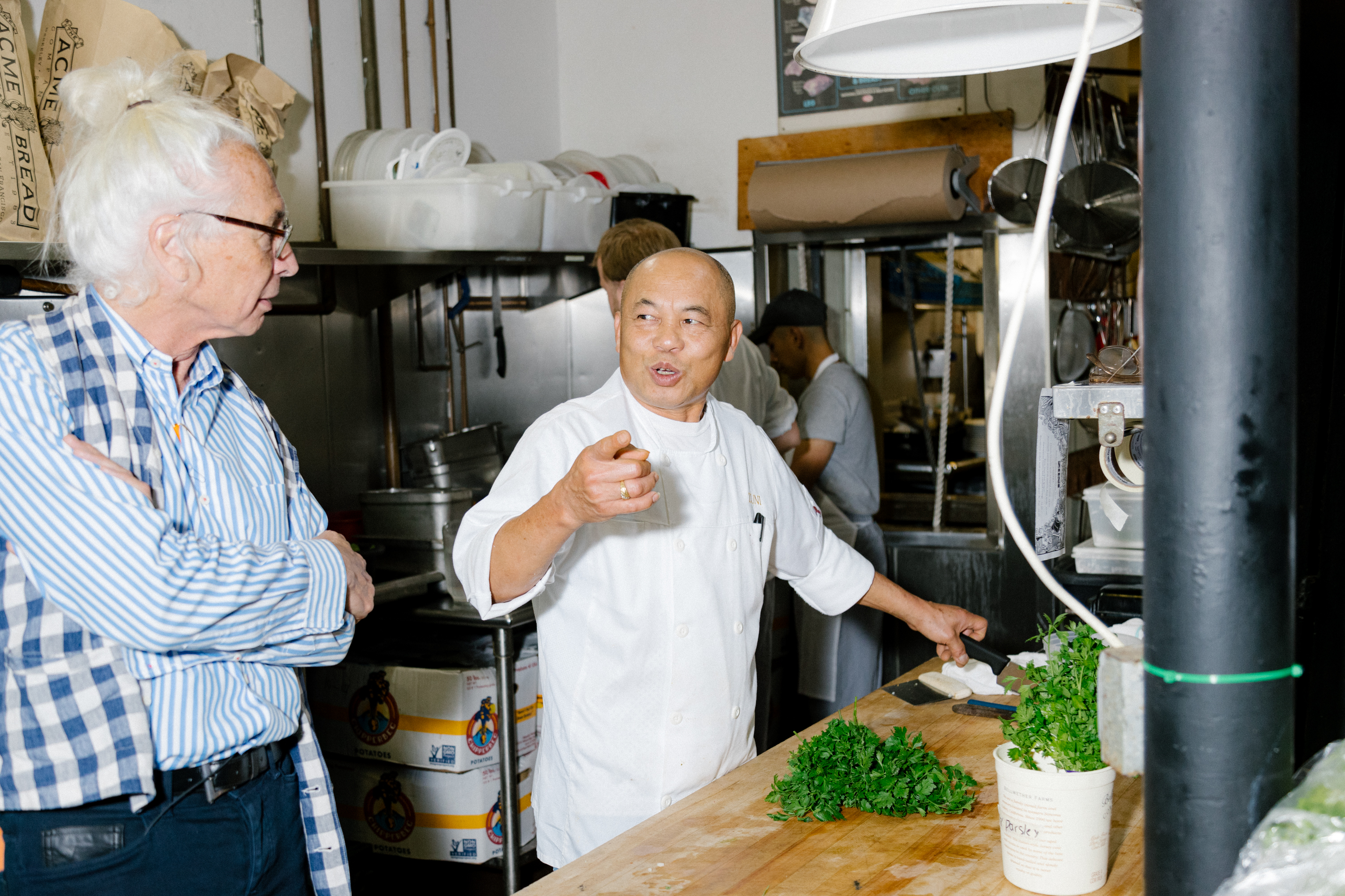 A chef speaks to a person in a kitchen area with parsely on the cutting table.