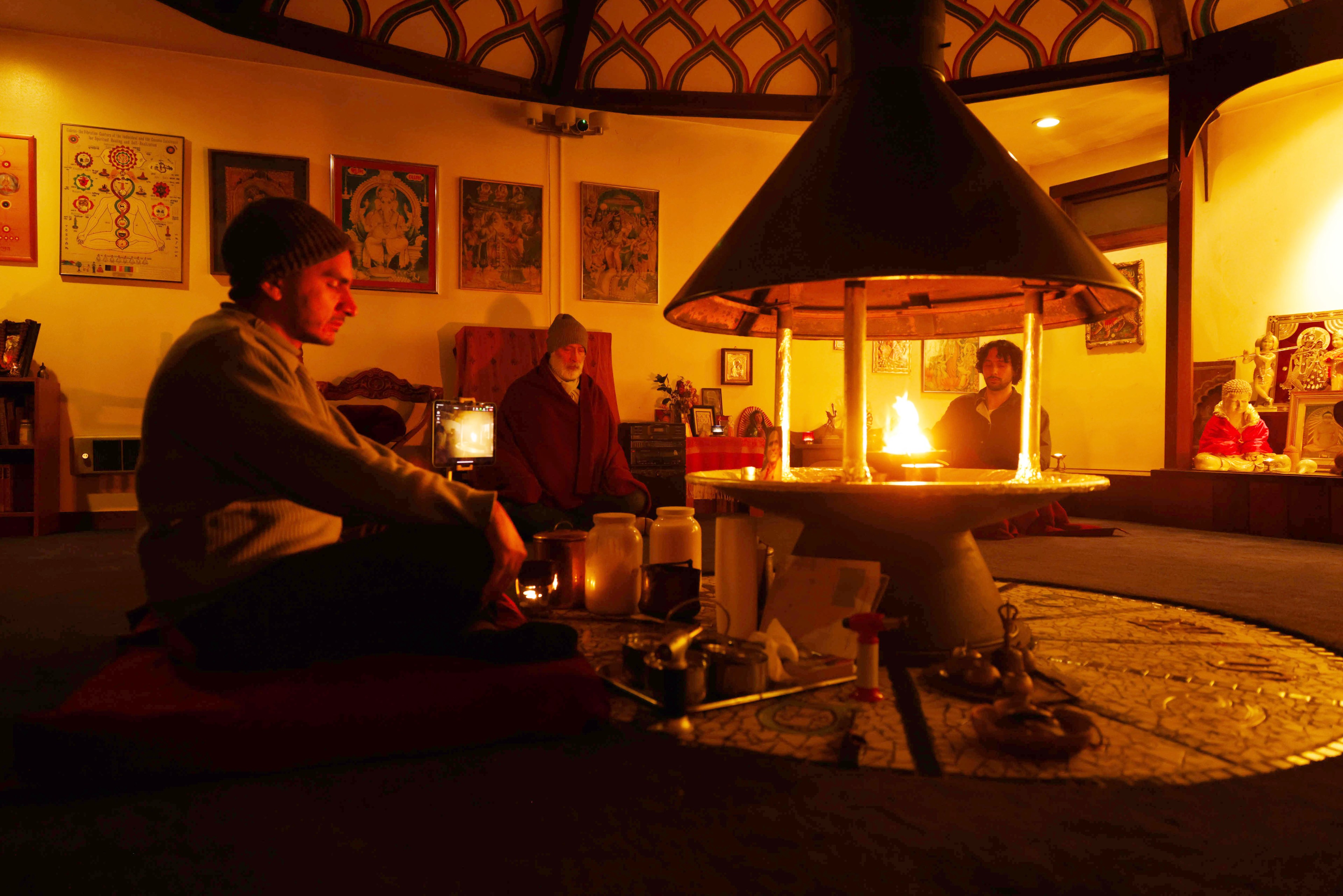 Three people sit arond a fire ceremony in the center of a room with Hindu gods and goddesses on the wall.