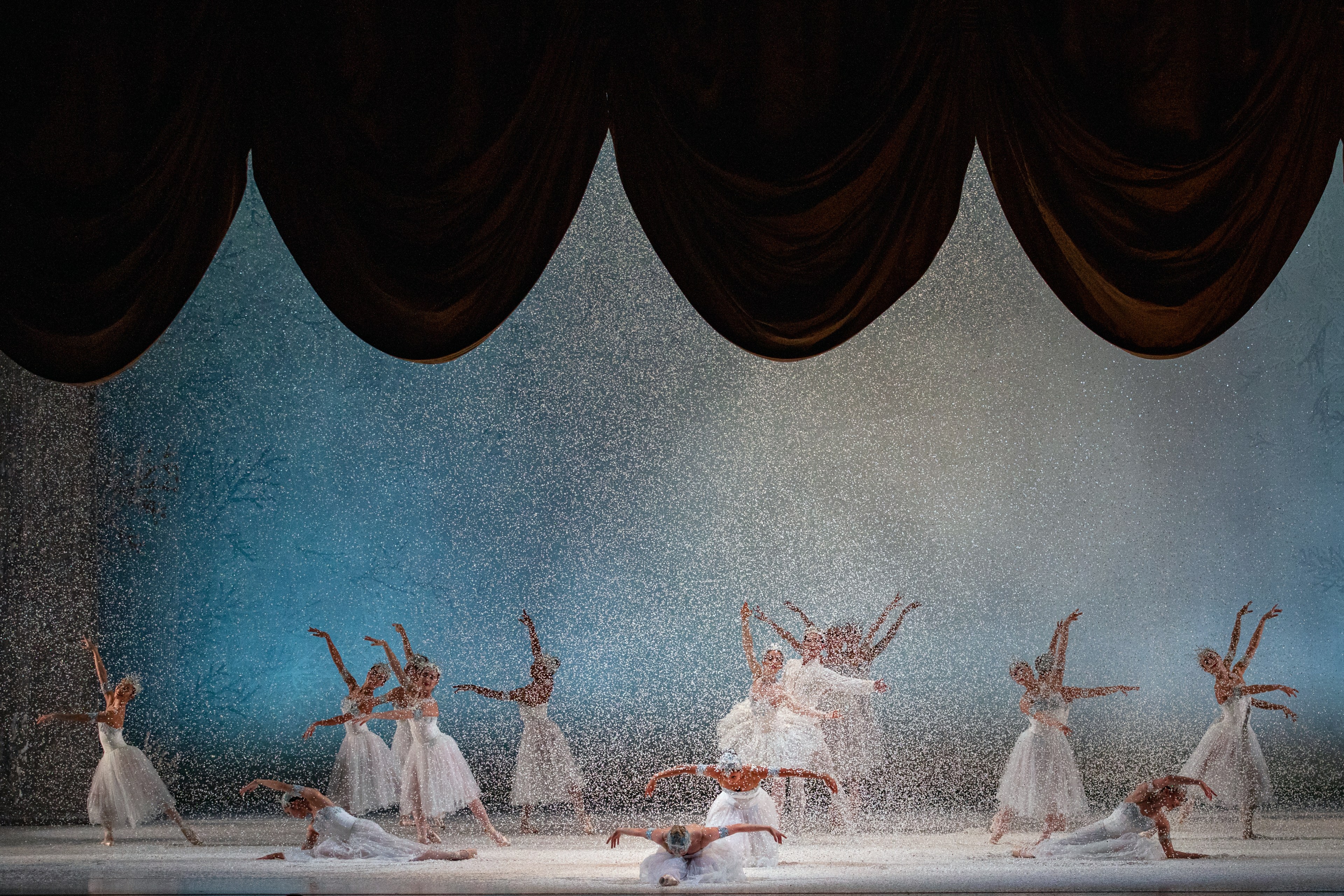 Ballet dancers perform on stage as snow falls with a large curtain closing above them.