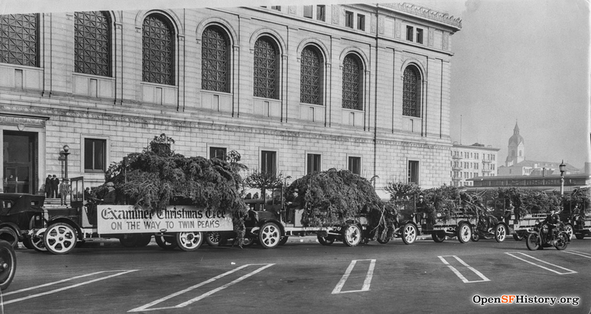 Trucks loaded with sections of a tree are seen in a caravan outside a neoclassical library.