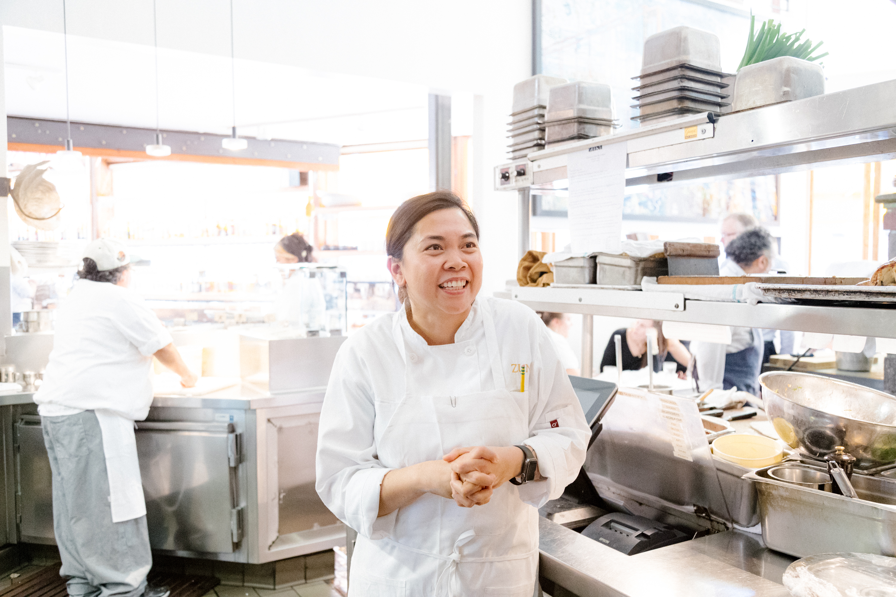 A chef smiles whiles clasping her hands in the back on an active kitchen.