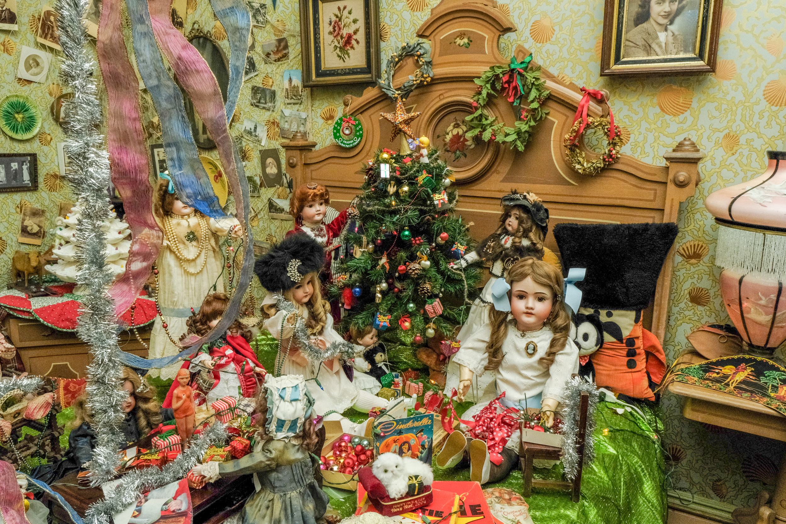 An antique wooden bed has a collection of antique objects on it, including vintage dolls and Christmas decorations.