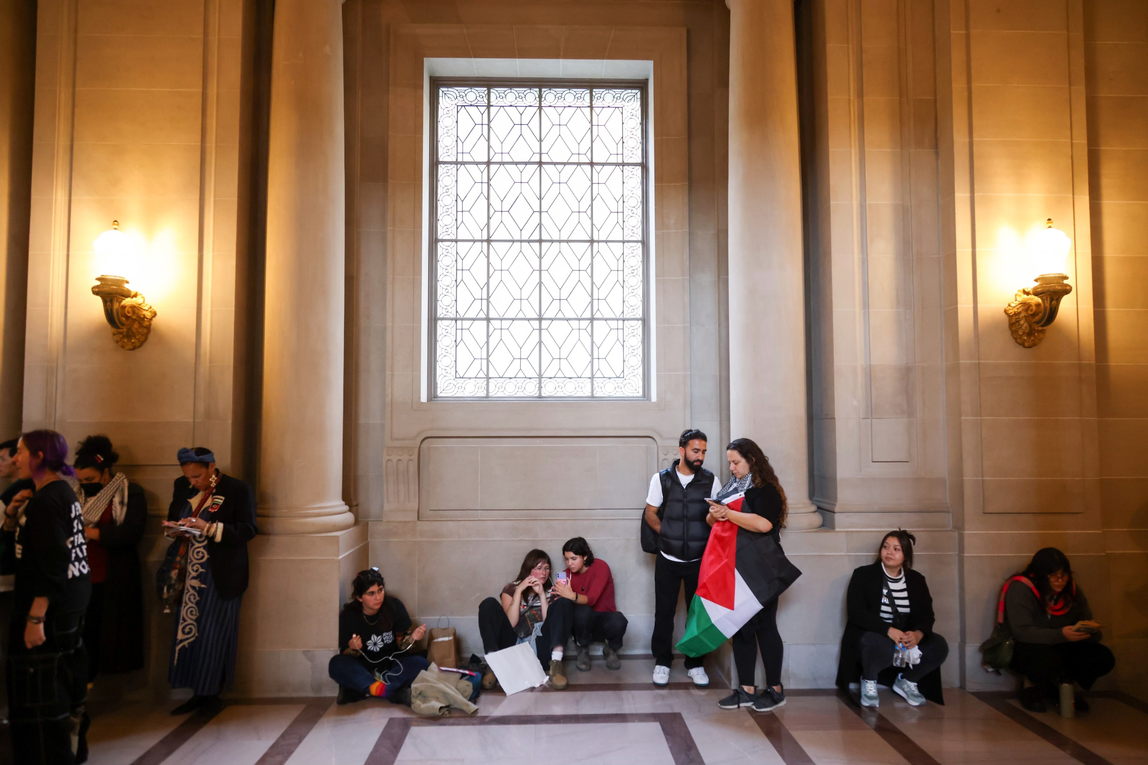 People sit and stand in a hall with tall walls, a large window, and ornate sconces. Two are holding a Palestinian flag.