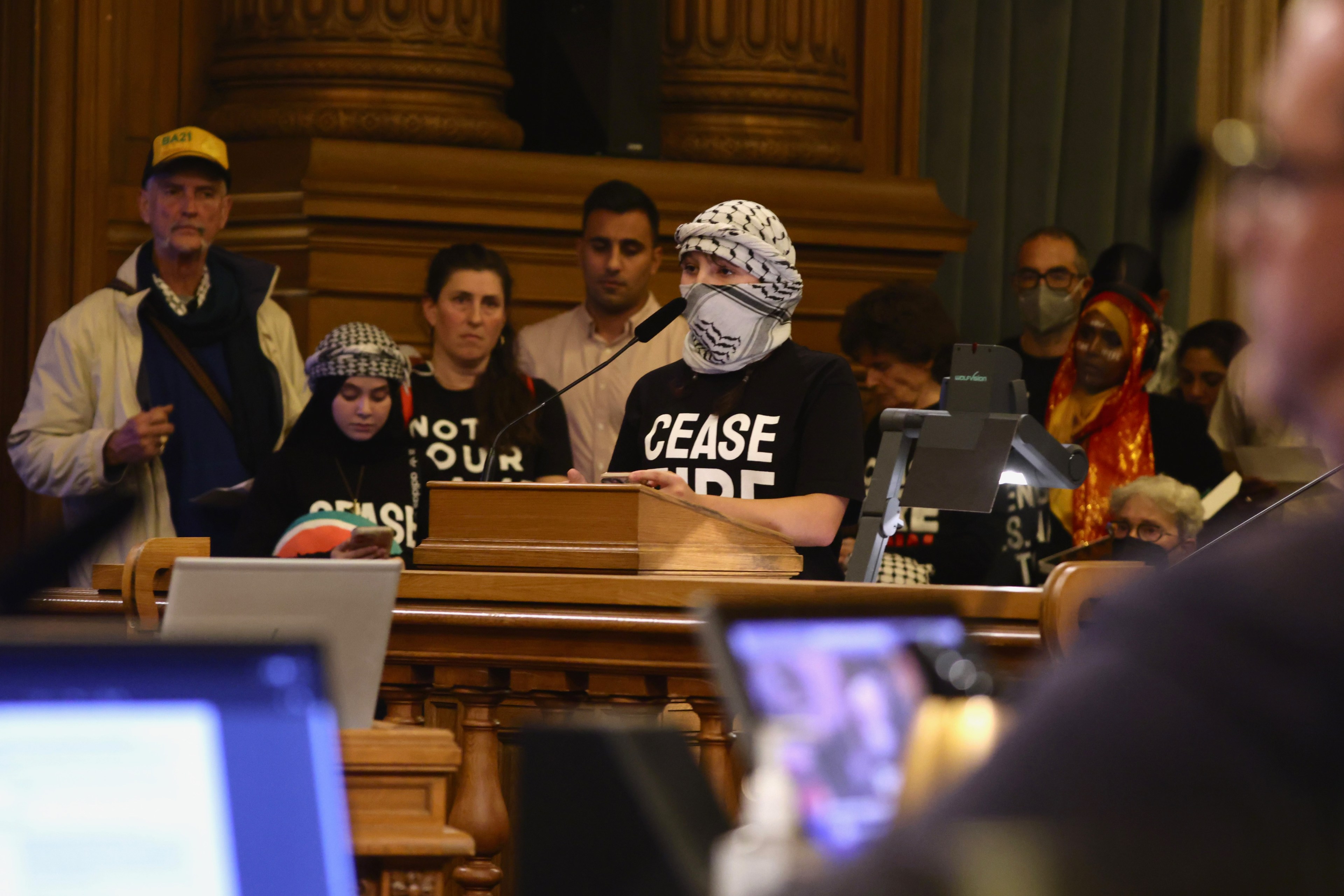 A person in a keffiyeh and mask speaks at a podium, with listeners wearing "Ceasefire" shirts behind them in a wood-paneled room.