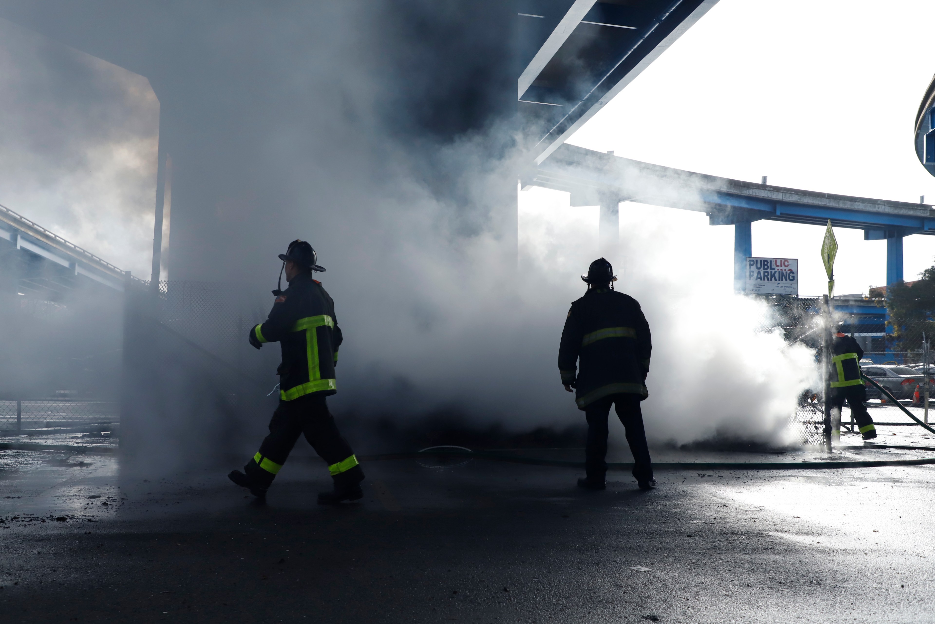 Firefighters walk through thick smoke, with hoses, near a "Public Parking" sign, under an overpass.