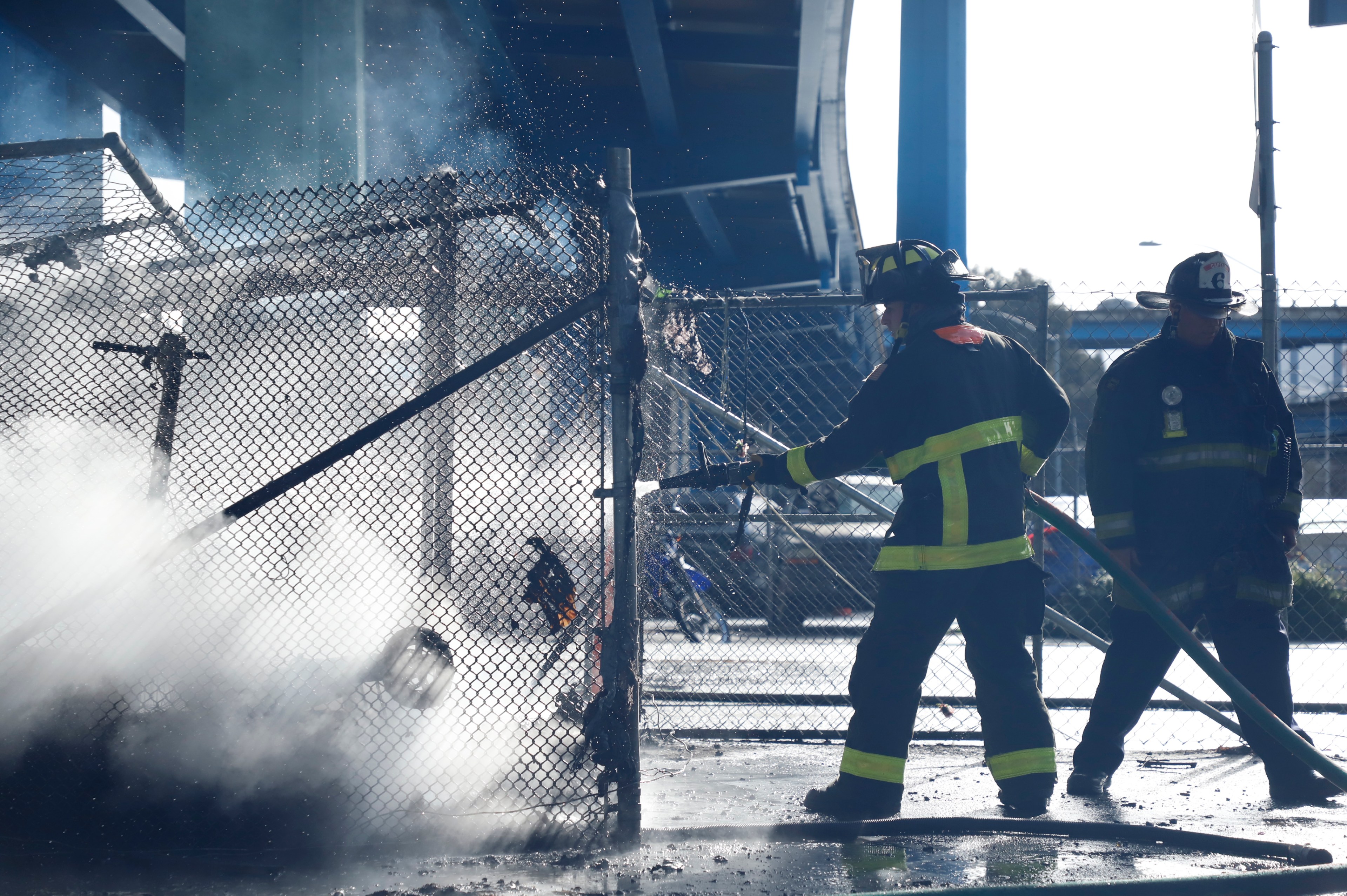 Two firefighters extinguish a fire behind a chain-link fence, water spraying through the air, under sunlight.