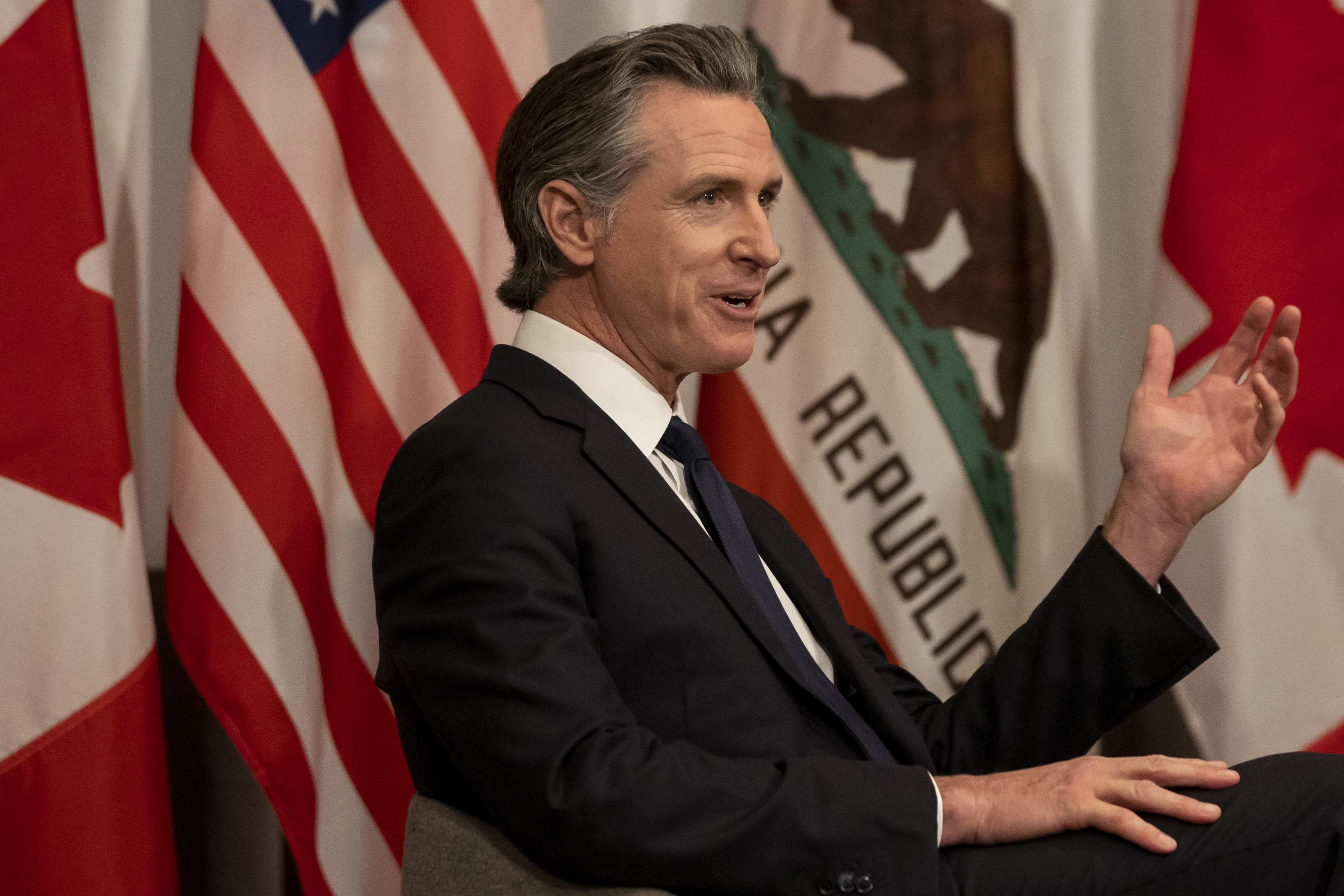 A man in a suit gestures while speaking, flanked by the US and California flags.
