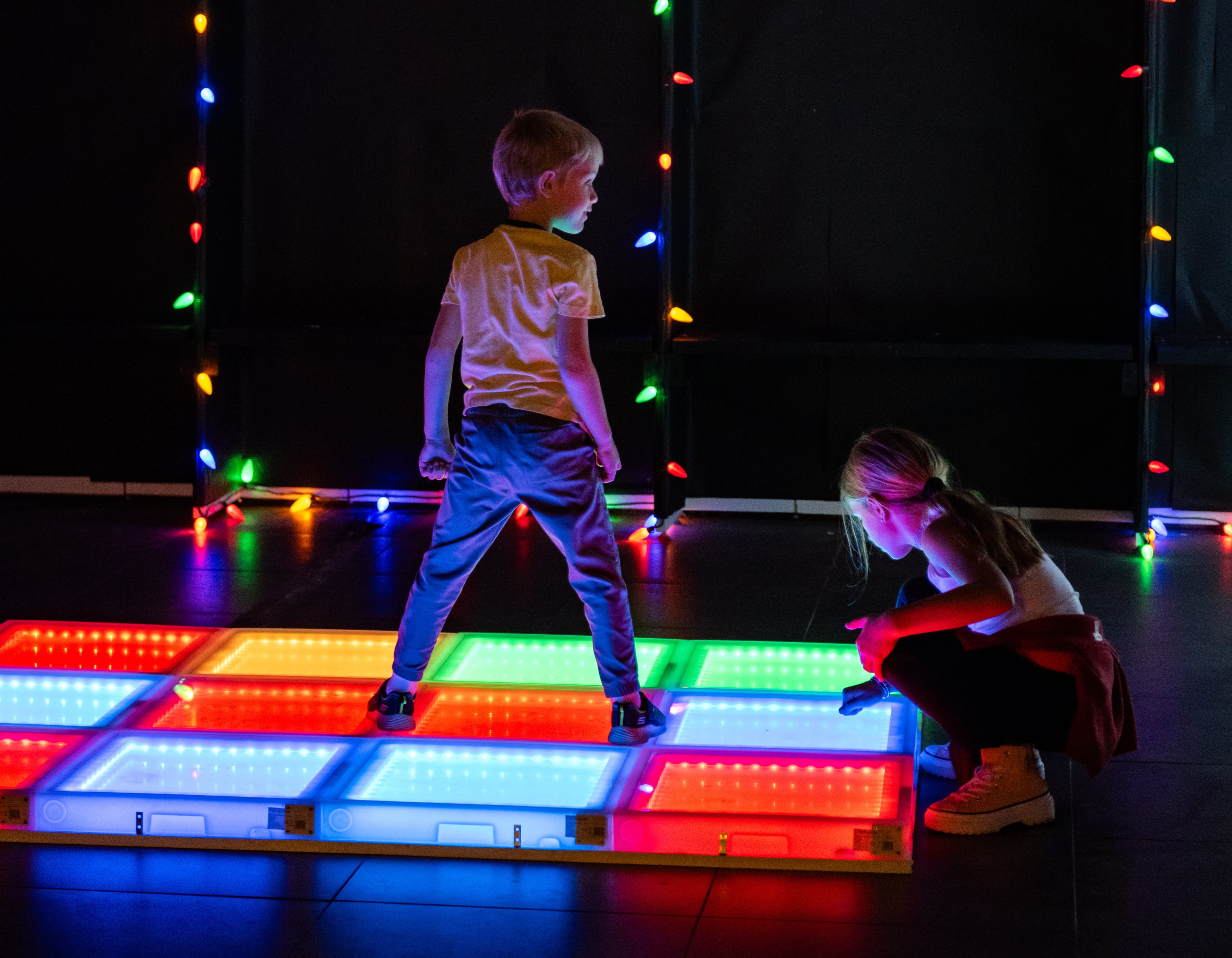 A little boy and girl play with an rainbow LED dance floor in a dark room decorated with lights for the holidays. 