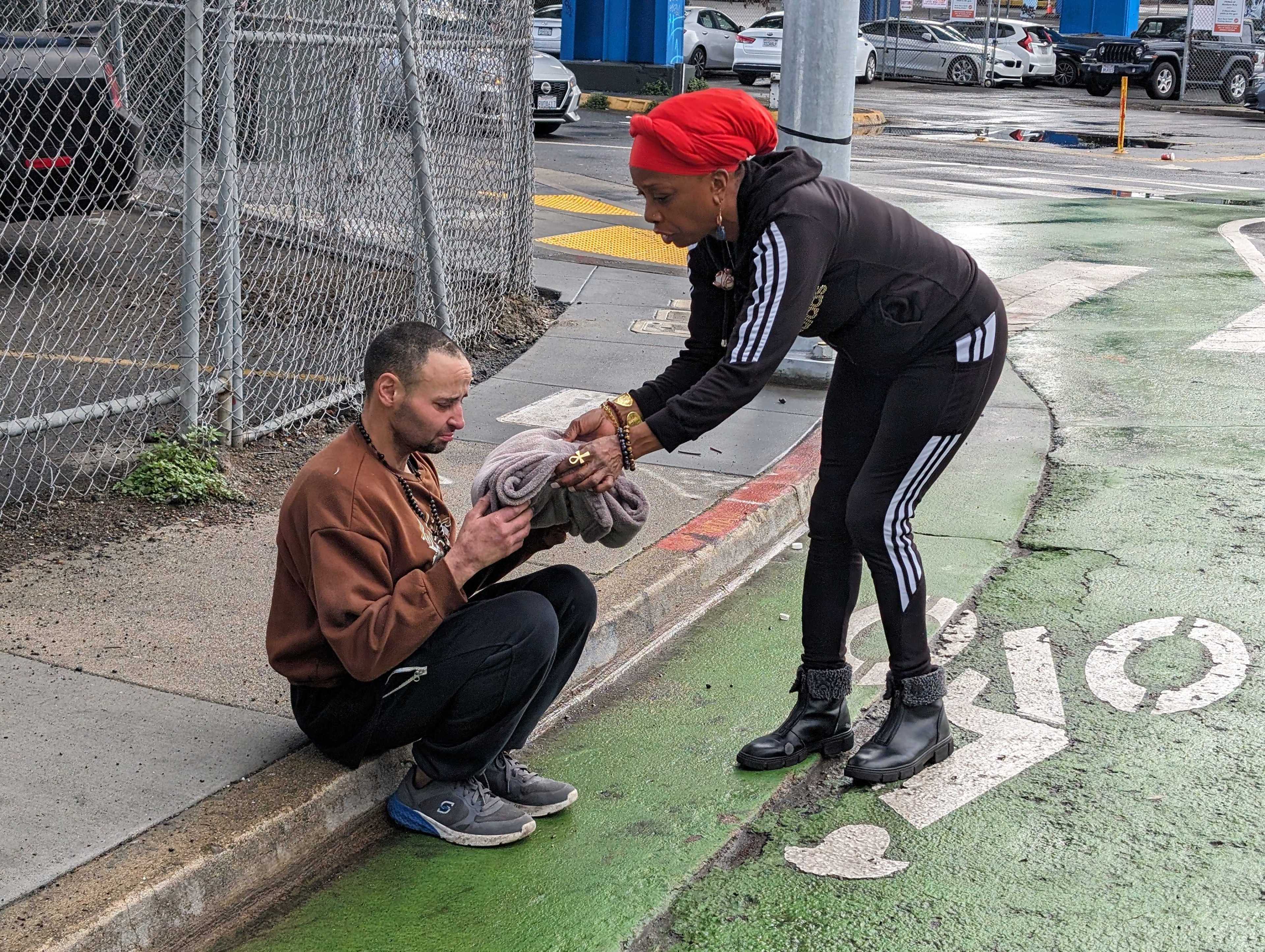 woman hands sobbing man a blanket as he sits on curb