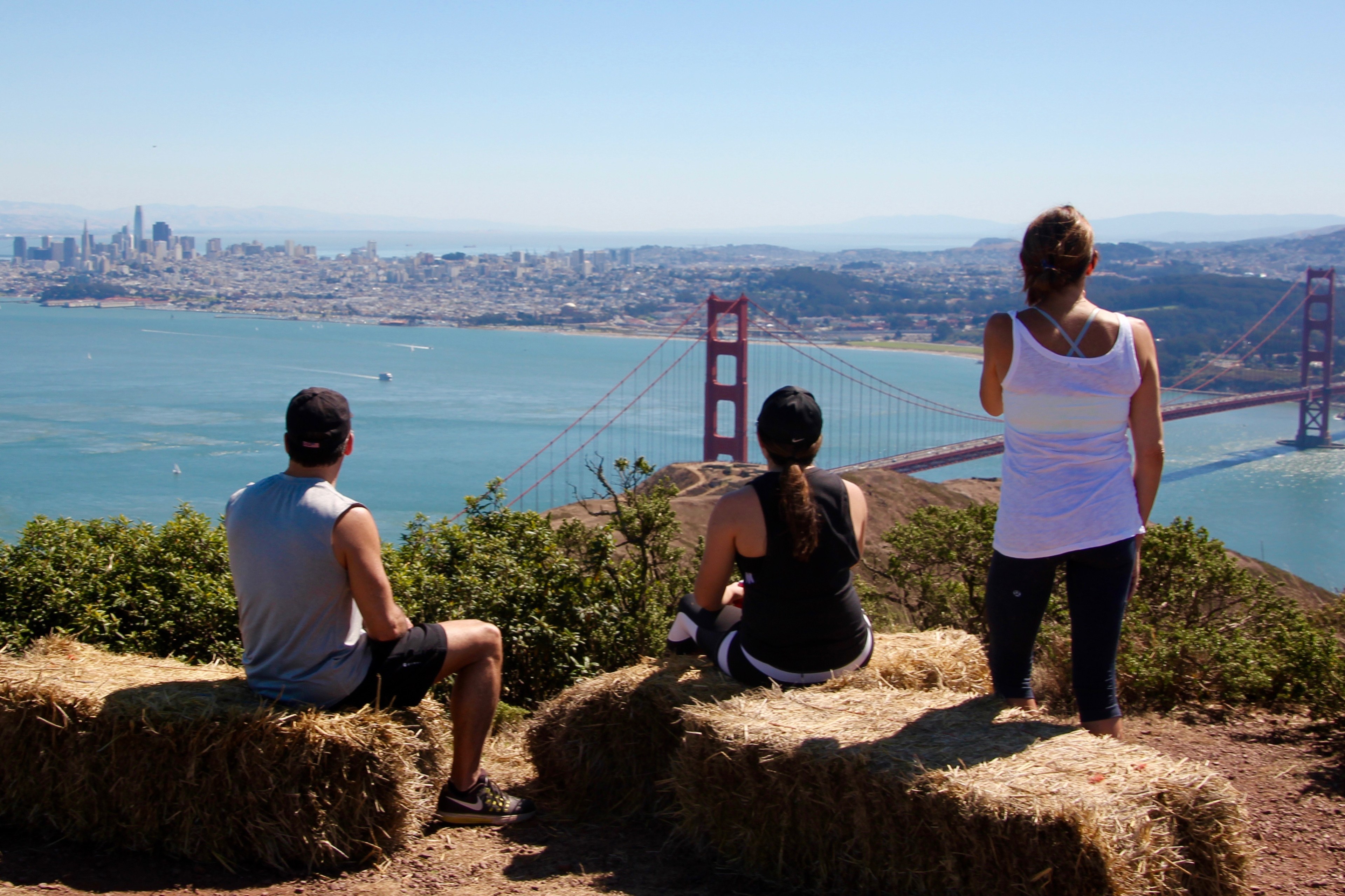 Three people looking at the Golden Gate Bridge and San Francisco skyline from a hill with hay bales.