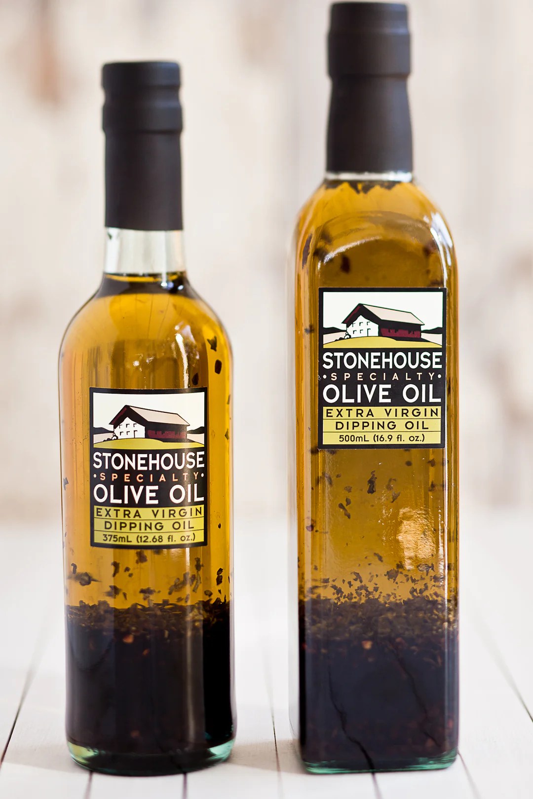 Two glass bottles of balsamic, herb dipping oil stand side by side with a blurred white background behind them.