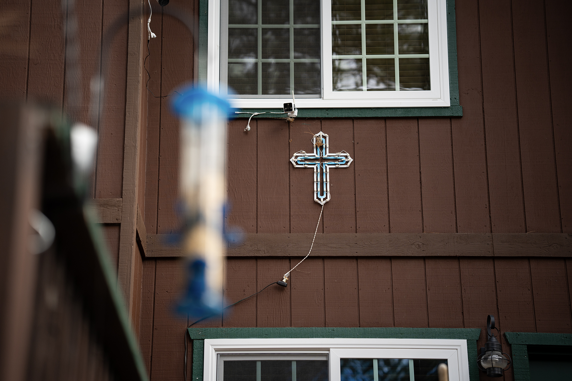 A blue and white cross light hands from the exterior of a brown house with a green trim around white windows