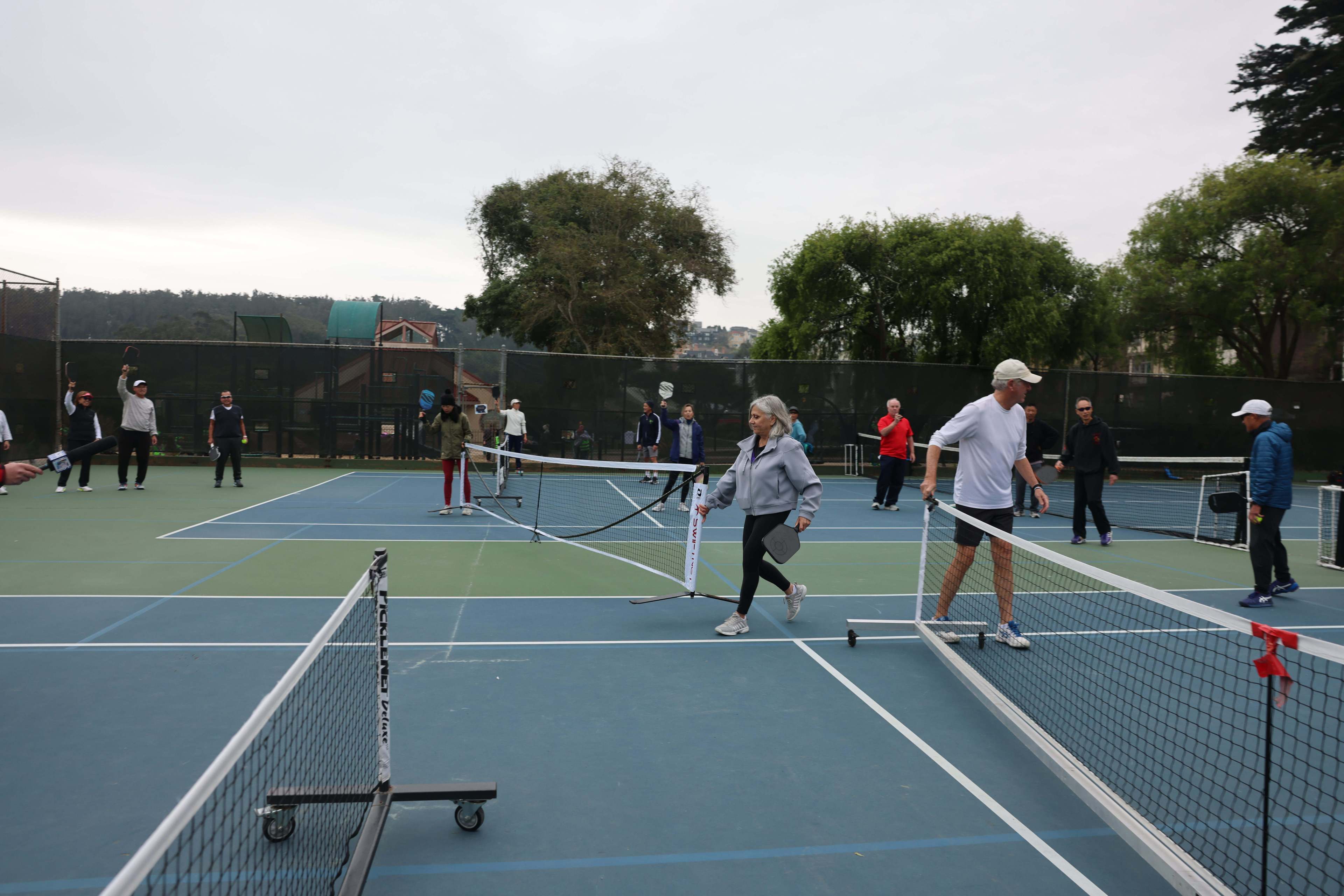 People playing tennis on multiple courts with trees and overcast sky in the background.