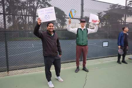 Two men stand on a tennis court, holding signs with "Make Pickleball Less Excruciating" and "FUN!" written on them.