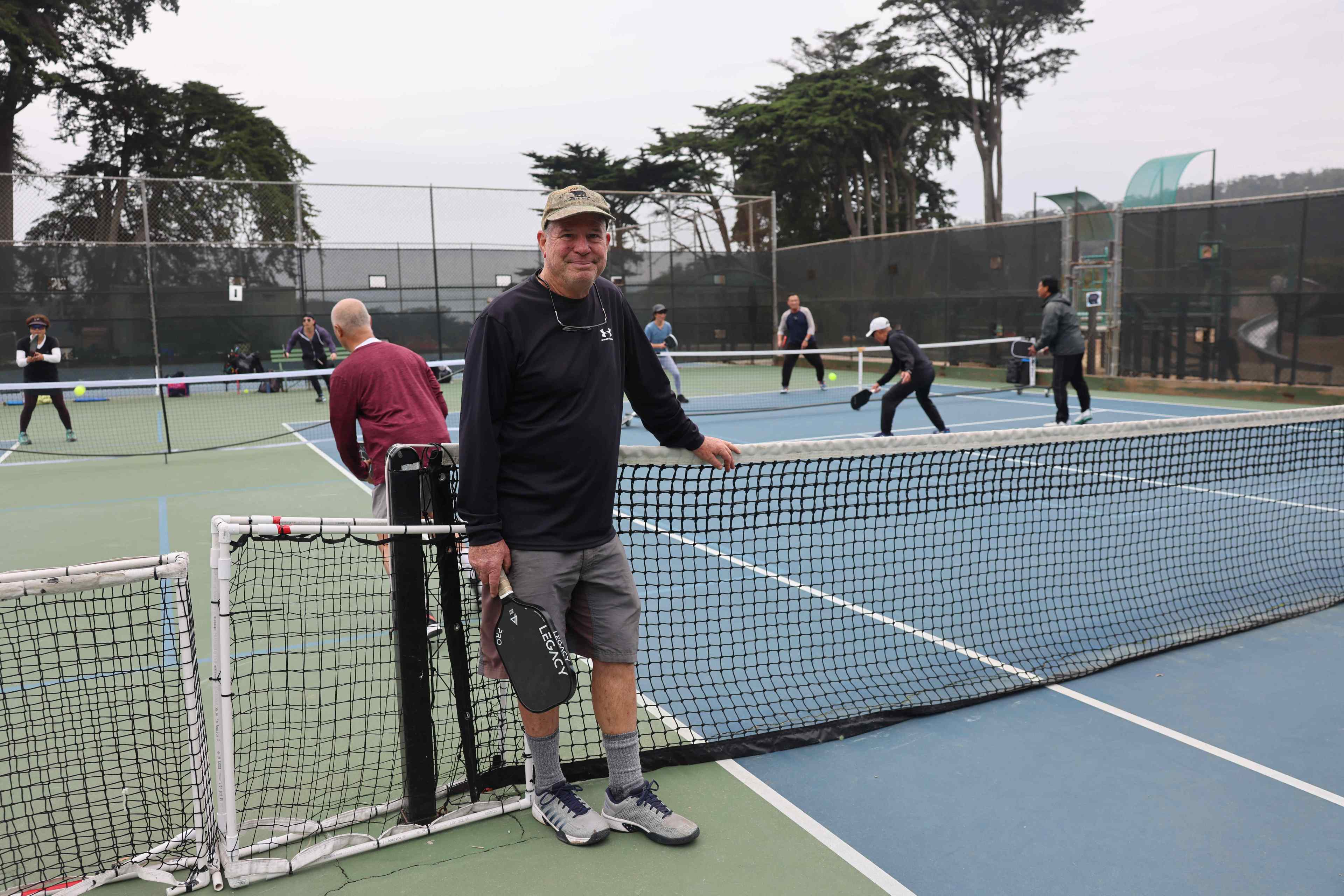A man stands smiling by a pickleball net, holding a paddle, with others playing in the background on the court.